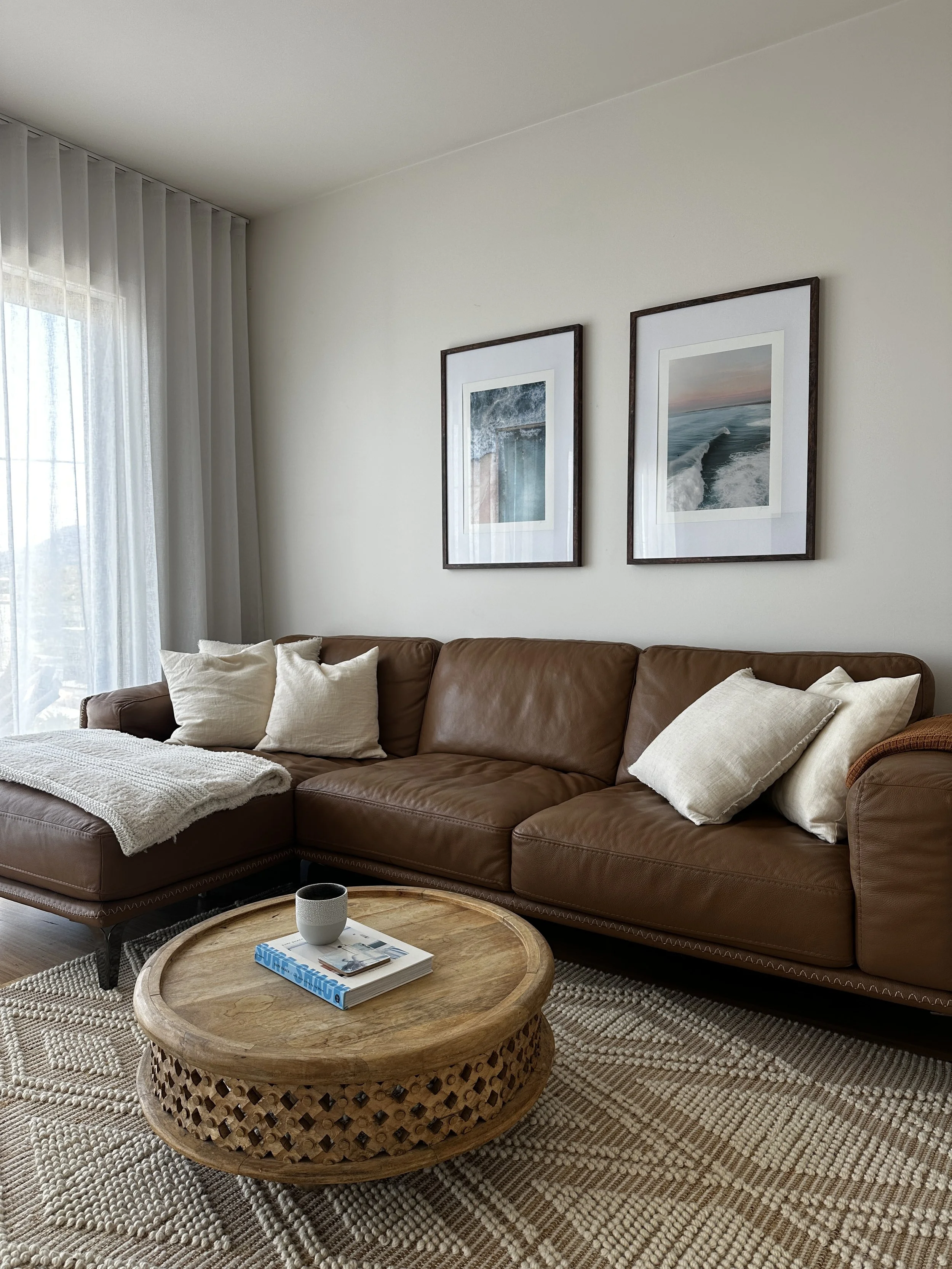 Living room with a brown leather sofa, beige pillows, a wooden coffee table with a magazine and a mug, a woven rug, and framed ocean-themed artwork on a white wall near a window with sheer curtains.