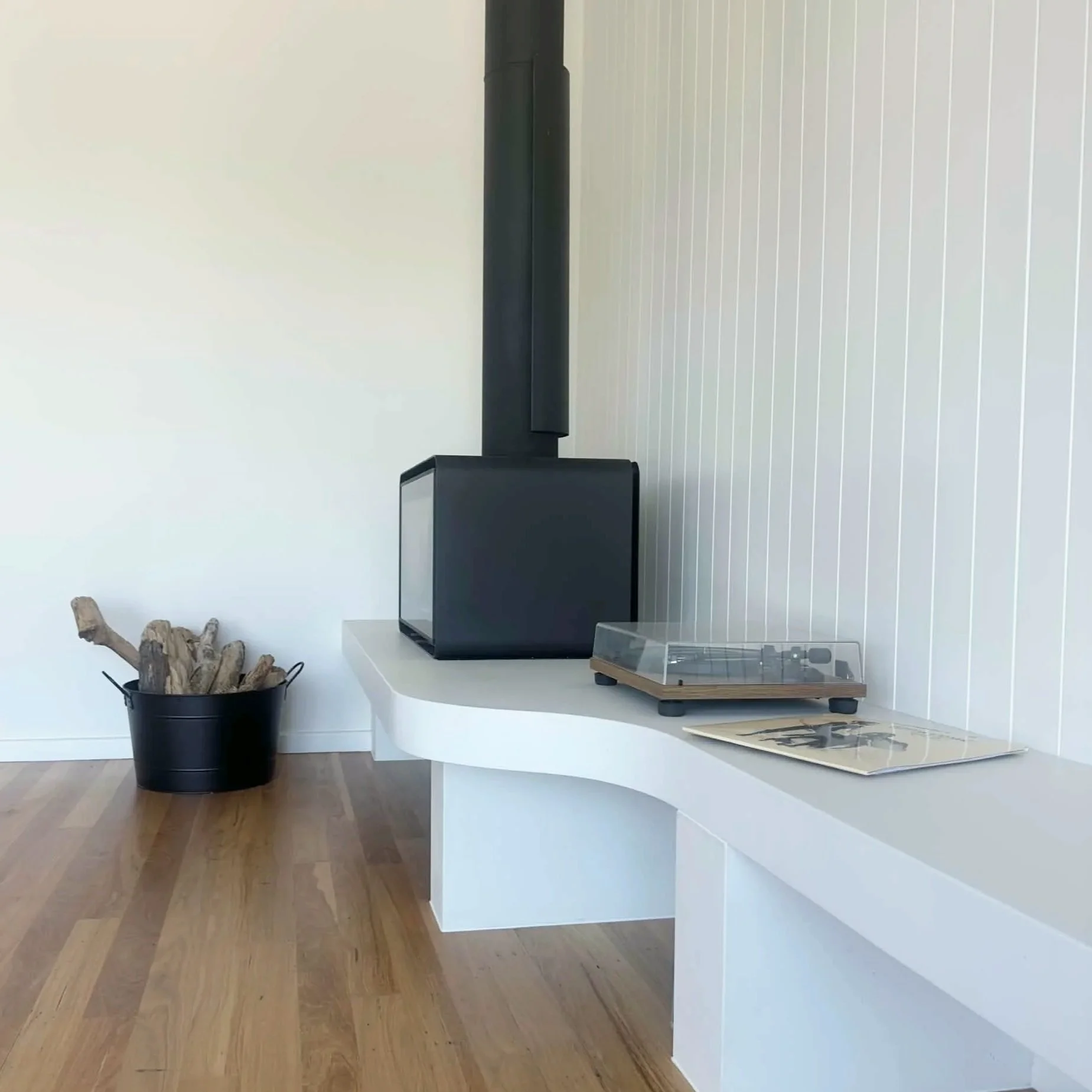 Interior of a room featuring a white curved counter, a black trash can filled with firewood, a black speaker with a tall black pole behind it, a wooden magazine holder, and magazines on the counter, with a light-colored wall and wooden floor.