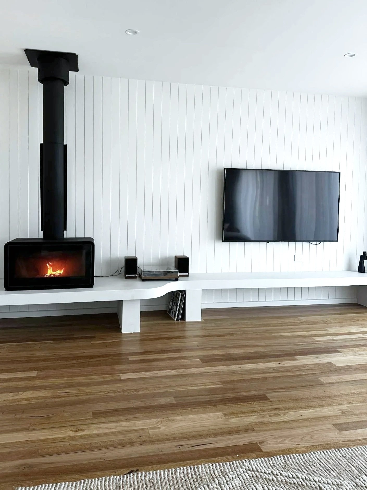 Living room with a flat-screen TV mounted on a white paneled wall, a wood stove with a fire burning inside, and wooden flooring with a light-colored rug.