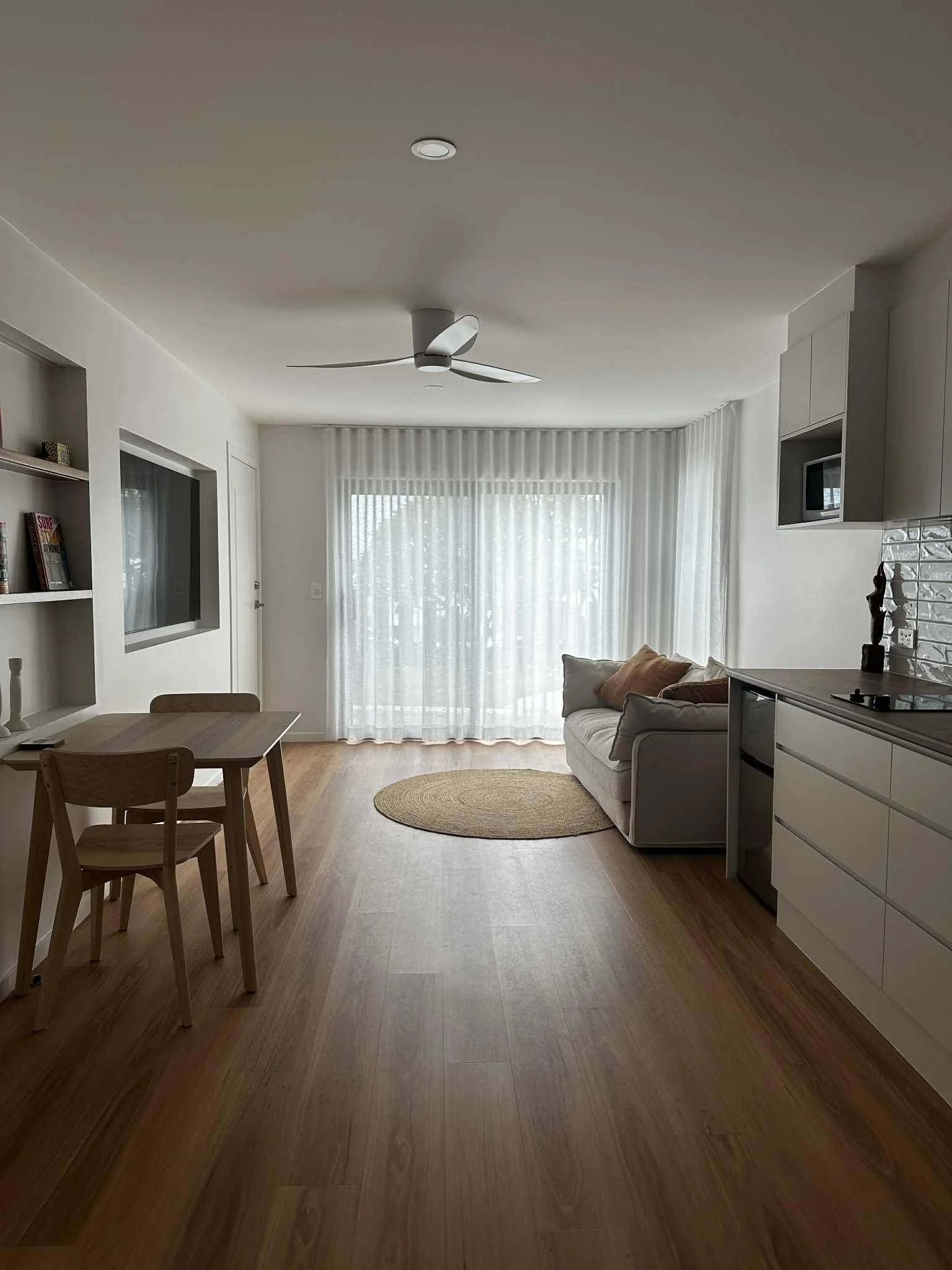 Living room with white walls, large window with sheer white curtains, white sofa with beige pillows, wooden floor, small oval rug, wall-mounted TV, ceiling fan, and kitchen area with white drawers and a microwave.
