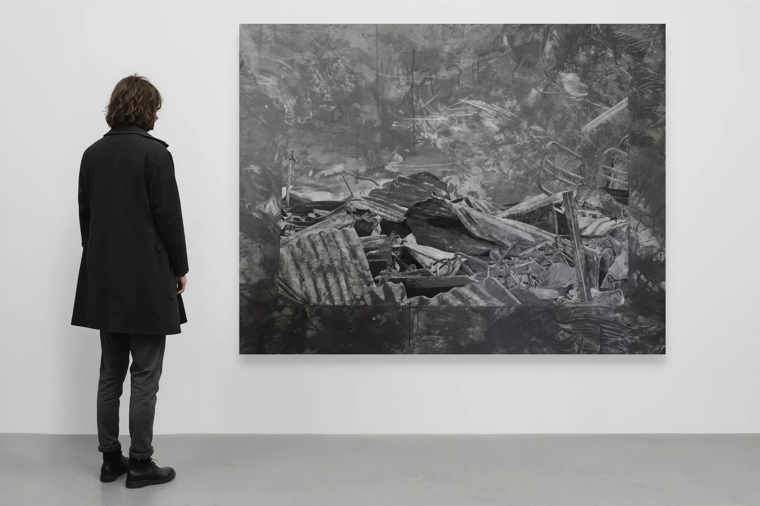 A person with shoulder-length hair, wearing a black coat and black boots, stands in front of a large black-and-white artwork depicting a chaotic scene of debris and wreckage. The setting appears to be an art gallery with white walls and a gray floor.