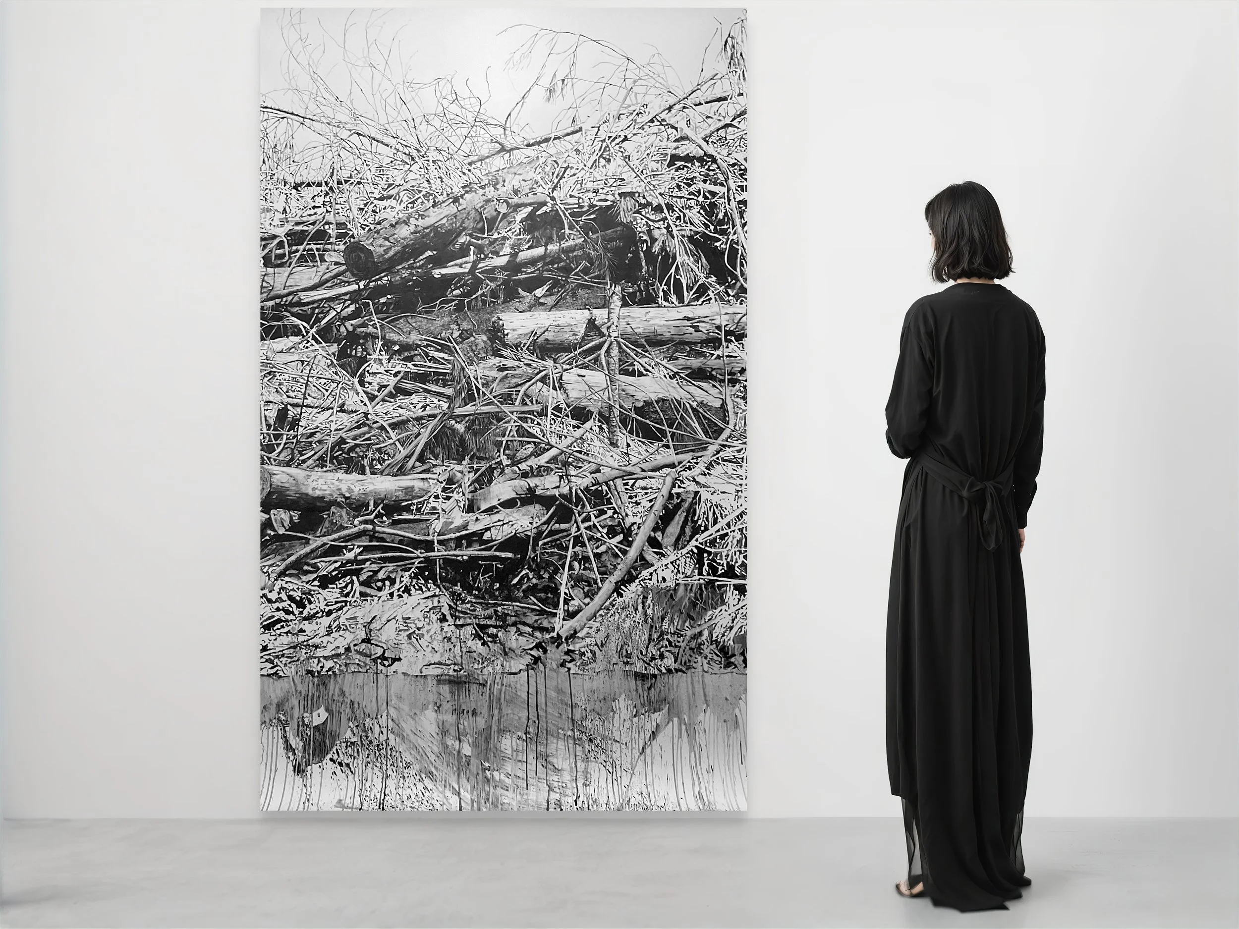 A woman with dark hair wearing a black dress observes a large black and white painting of fallen trees and branches on a white gallery wall.