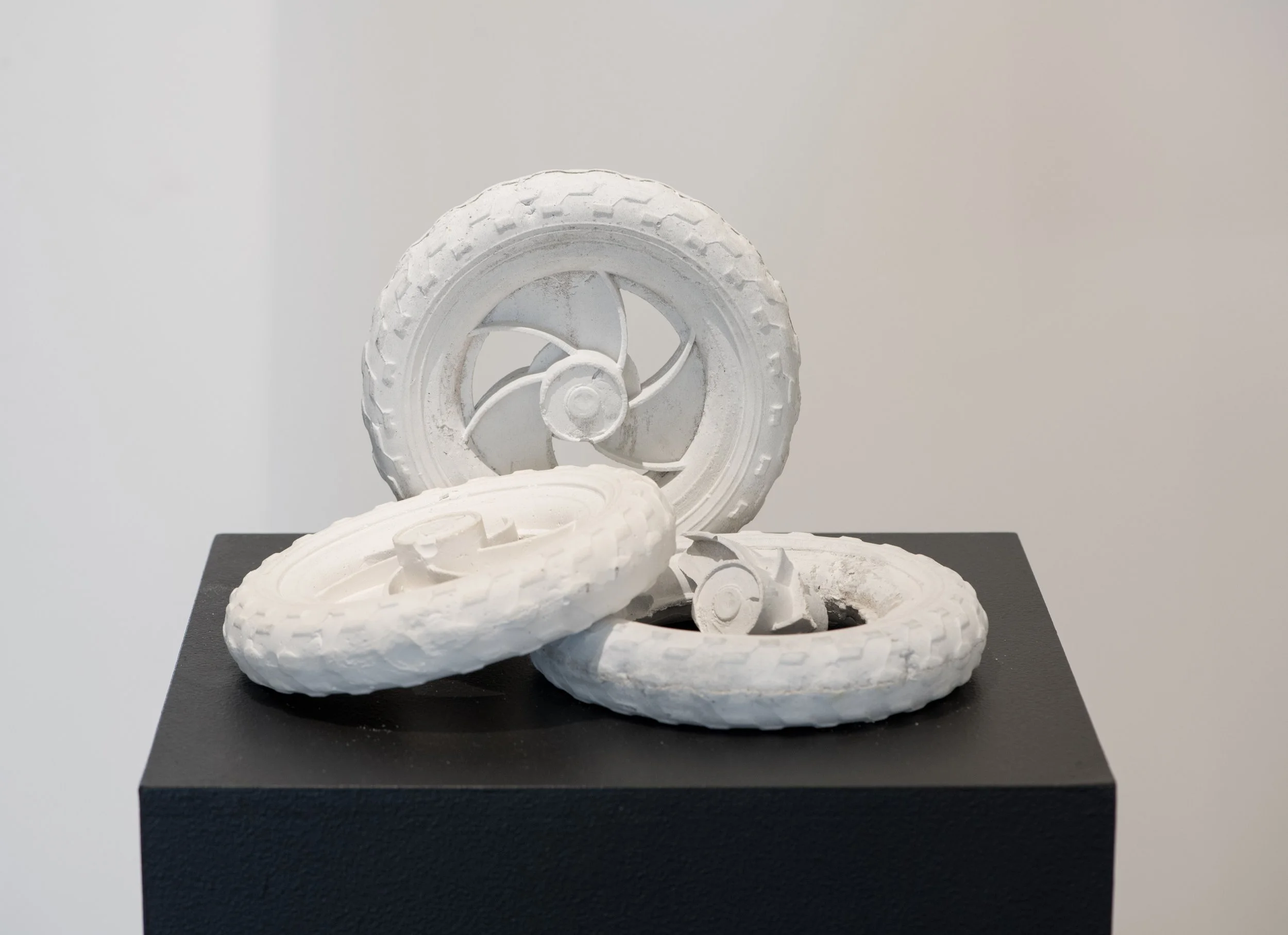 White sculptural artwork consisting of child's scooter wheels with decorative elements, displayed on a black base
