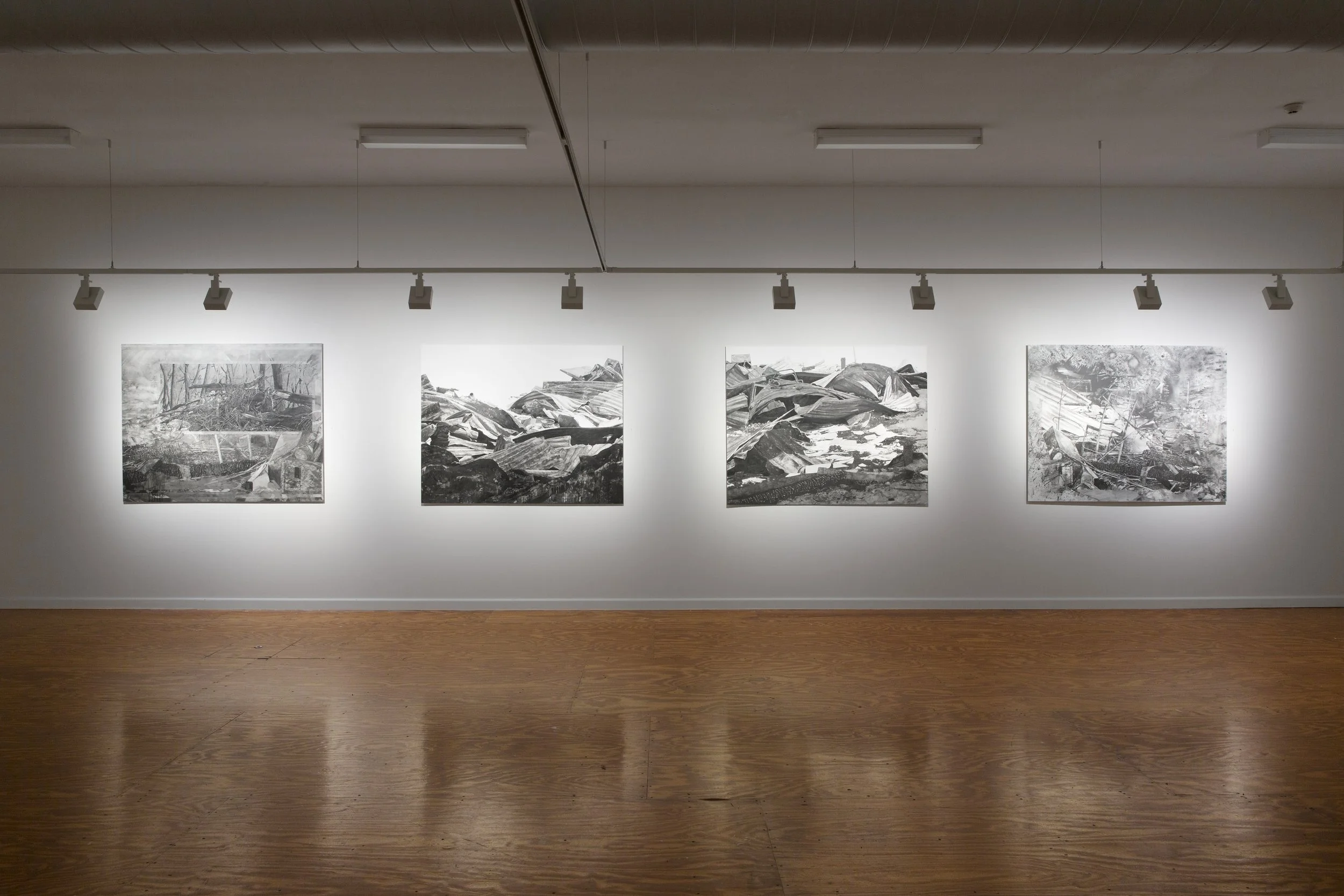 Black and white photographs of a destroyed landscape with debris and wreckage are displayed on a gallery wall in an art exhibit.