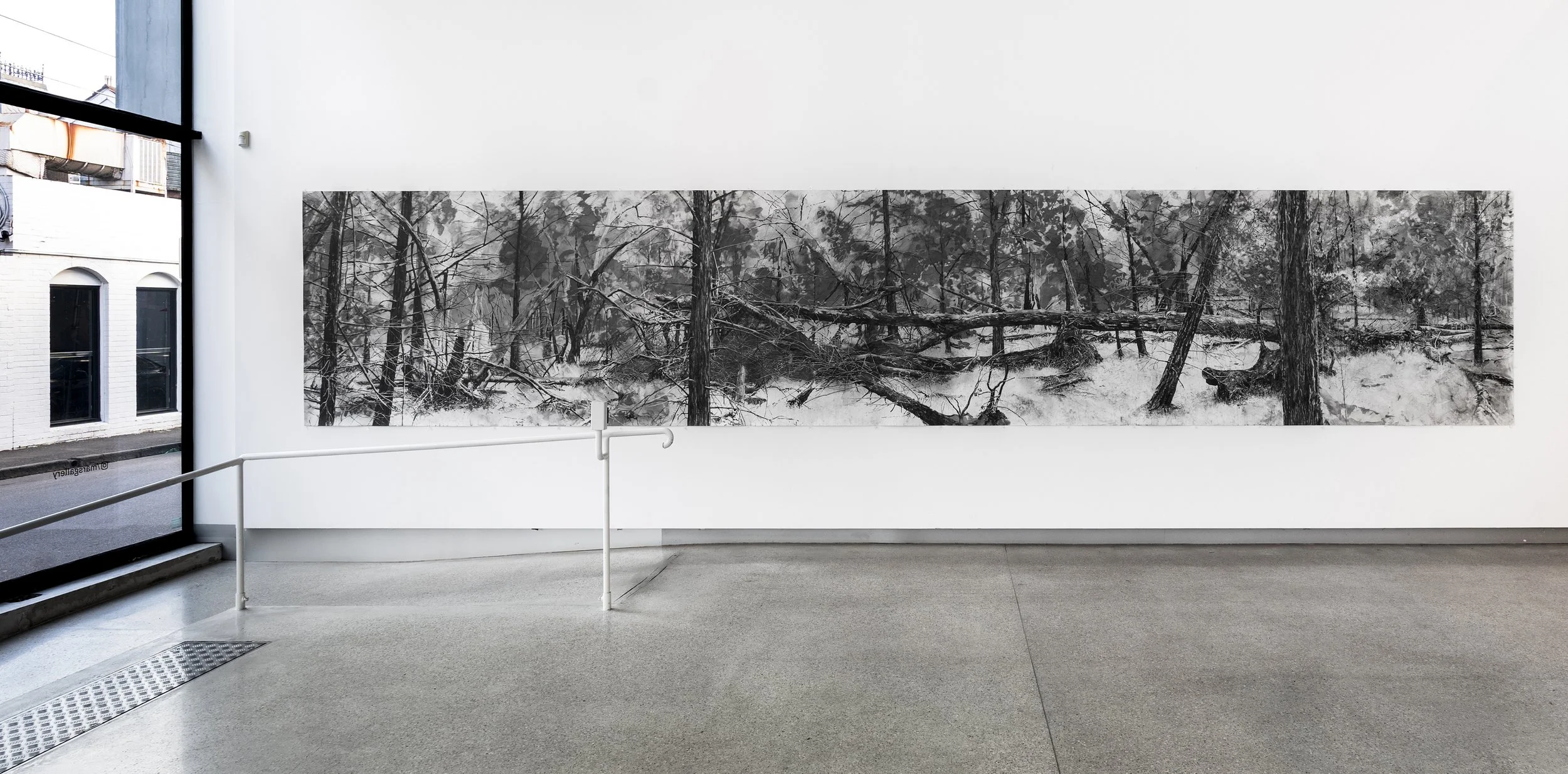 Black and white artwork of a forest scene with trees and fallen branches, displayed on a white gallery wall inside an art gallery.