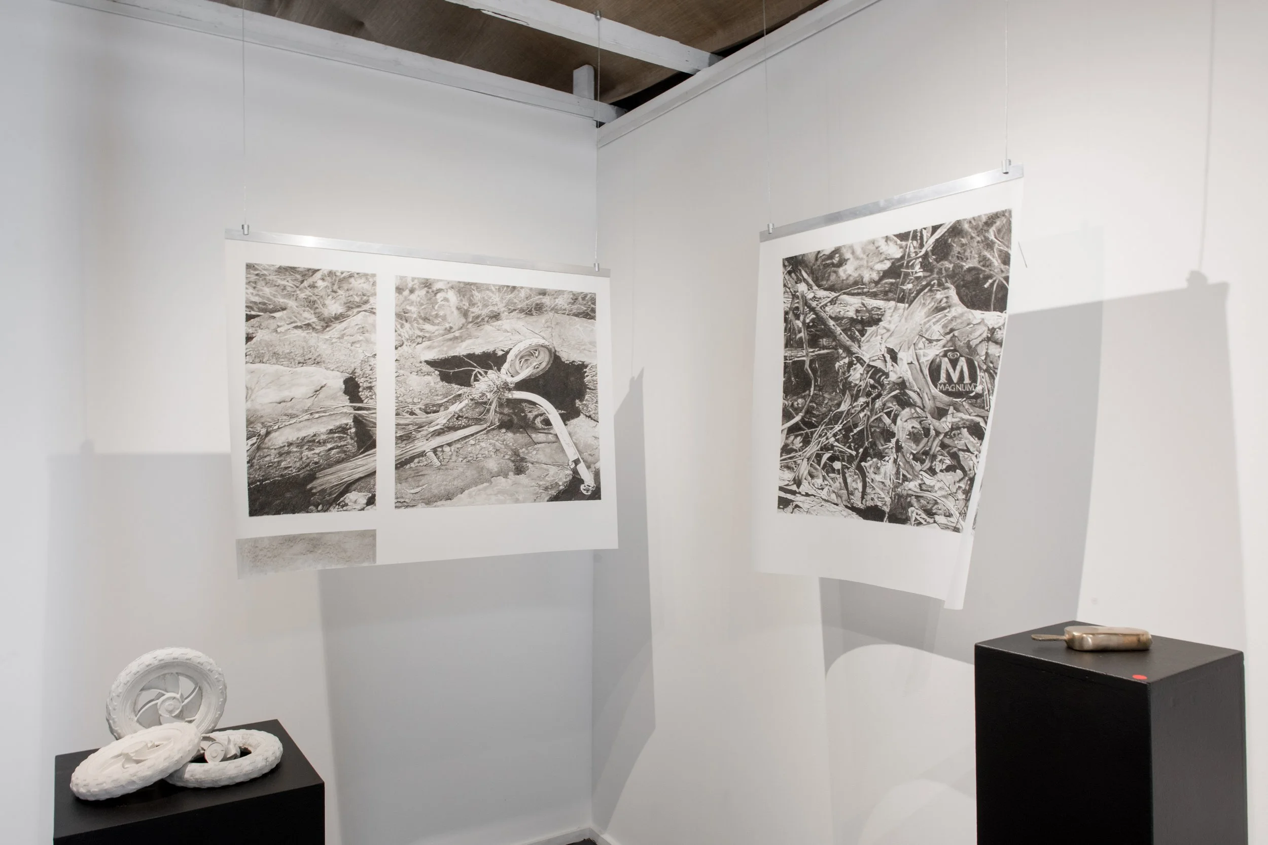 Art gallery display with black and white drawings hanging on white walls, and small white and bronze sculptures on black pedestals.