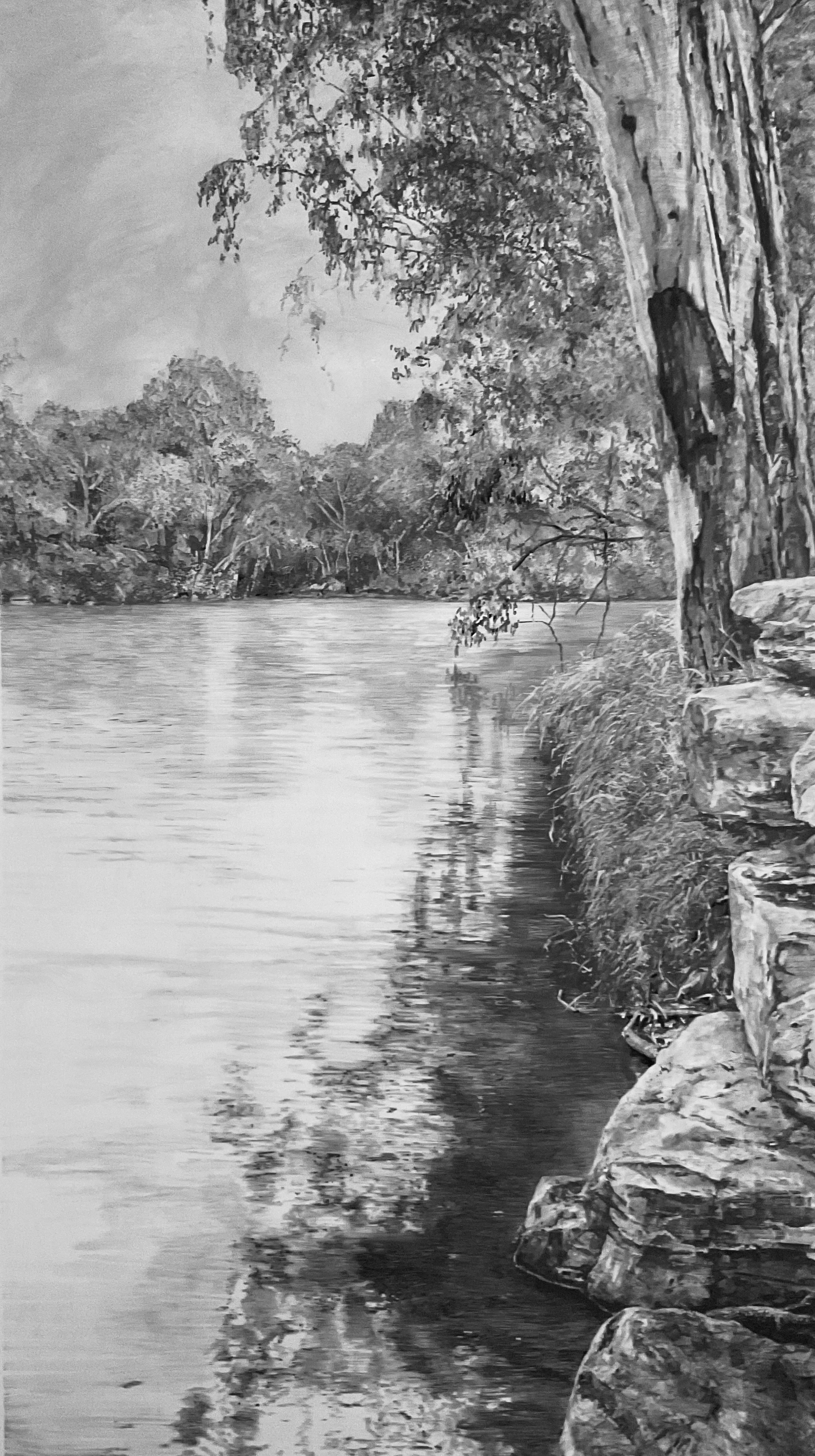A black and white photograph of a river with a rocky shoreline on the right and trees along the water's edge.