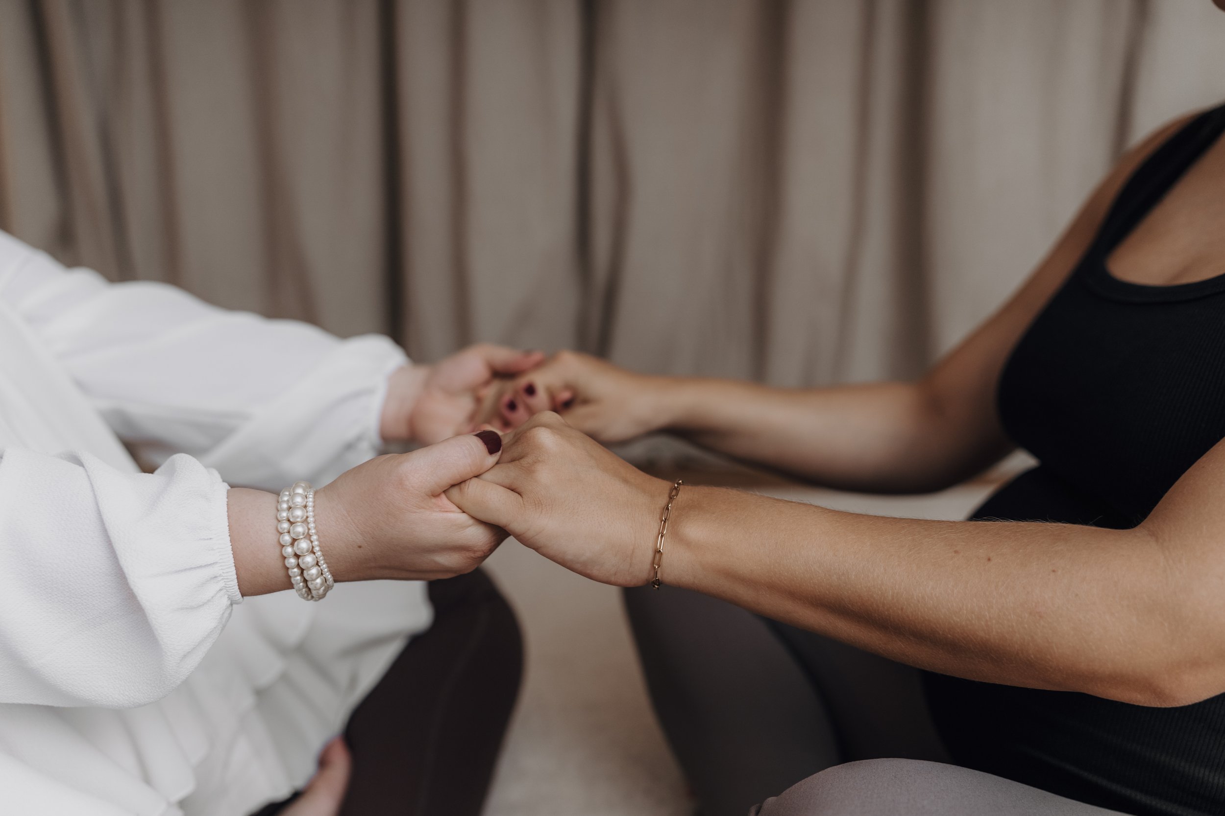 Zwei Frauen halten die Hände des anderen und berühren sich auf symbolische Weise bei einer emotionalen Begegnung, im Hintergrund ein Vorhang.