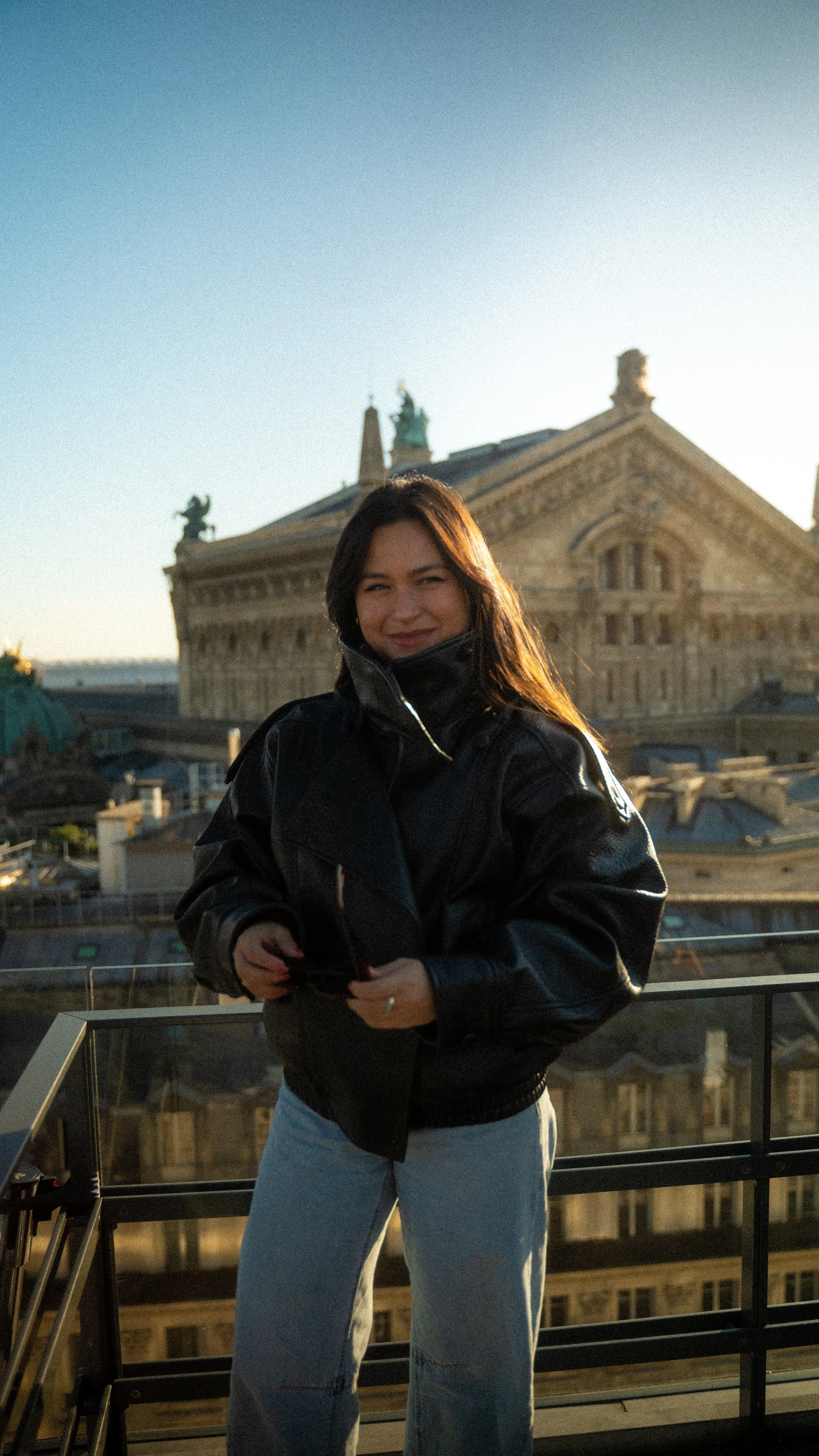 A woman with long dark hair smiling at sunset, wearing a black leather jacket and light blue jeans, standing on a rooftop with historic buildings behind her.