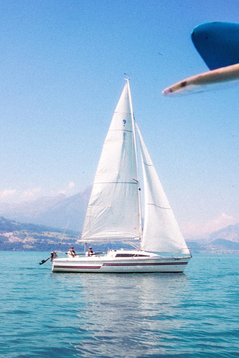 A sailboat with white sails on a calm body of water, with distant mountains and a blue sky in the background.