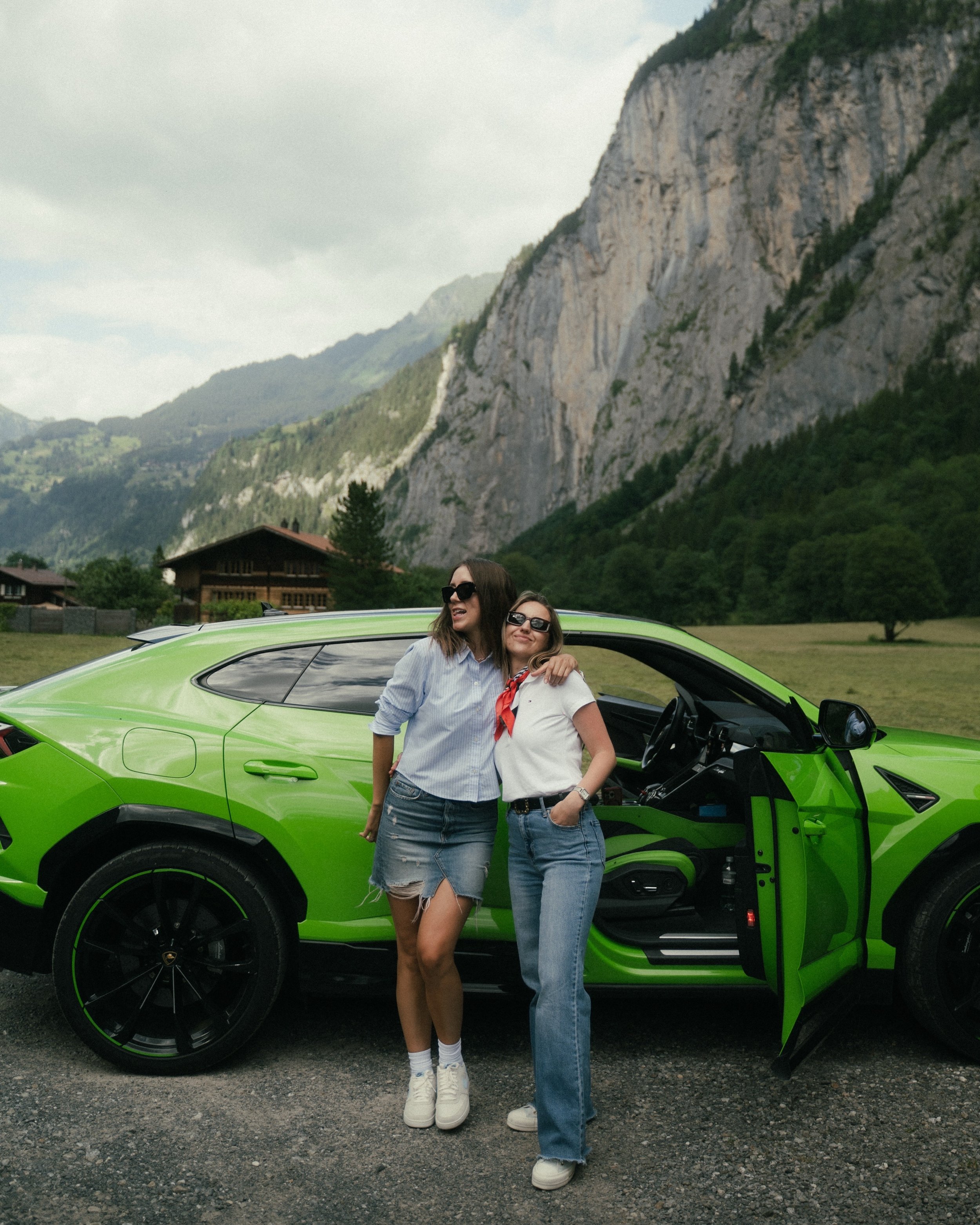 Two women standing outside next to a bright green Lamborghini with mountains in the background. They are smiling and wearing sunglasses.