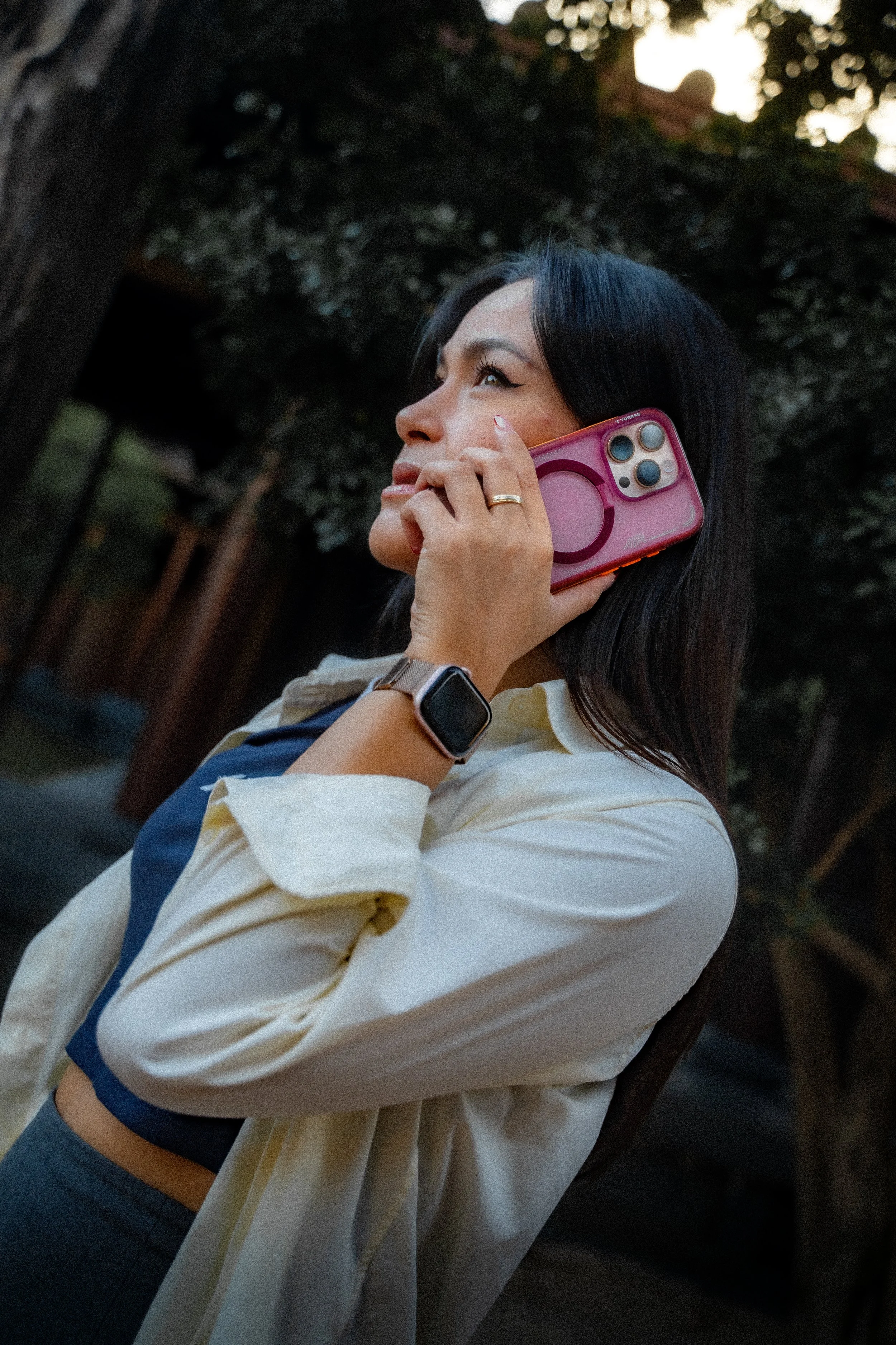 A woman with black hair and makeup talking on a pink smartphone outdoors during the evening or late afternoon. She wears a beige and navy shirt, gray pants, a watch, and a ring.