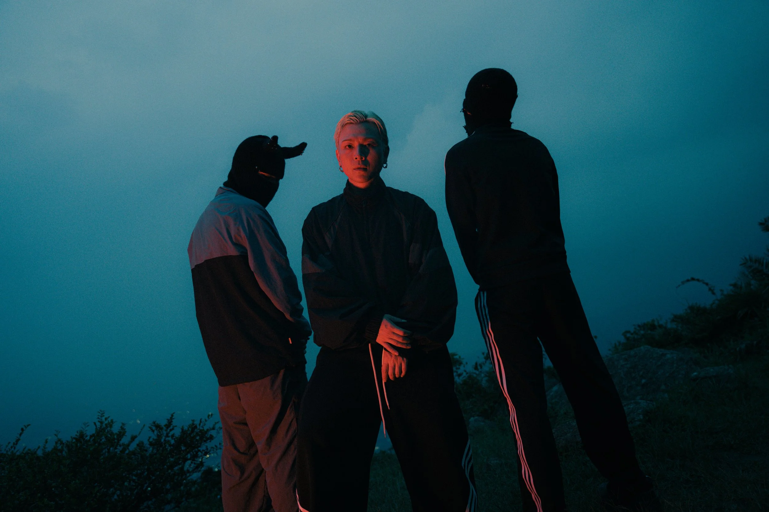 Three individuals standing outdoors during dusk or night, silhouetted against a blue sky, with some shrubbery visible at the bottom of the image. The person in the middle has blonde hair and light skin, while the others have darker skin and are weari