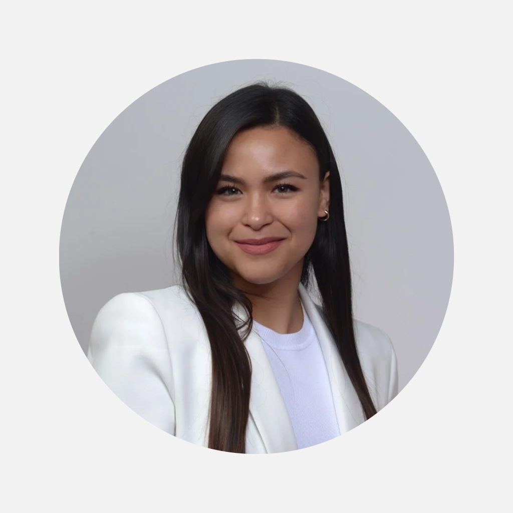 Une femme souriante avec des cheveux longs et noirs, portant un blazer blanc et une chemise blanche, posant contre un fond gris.