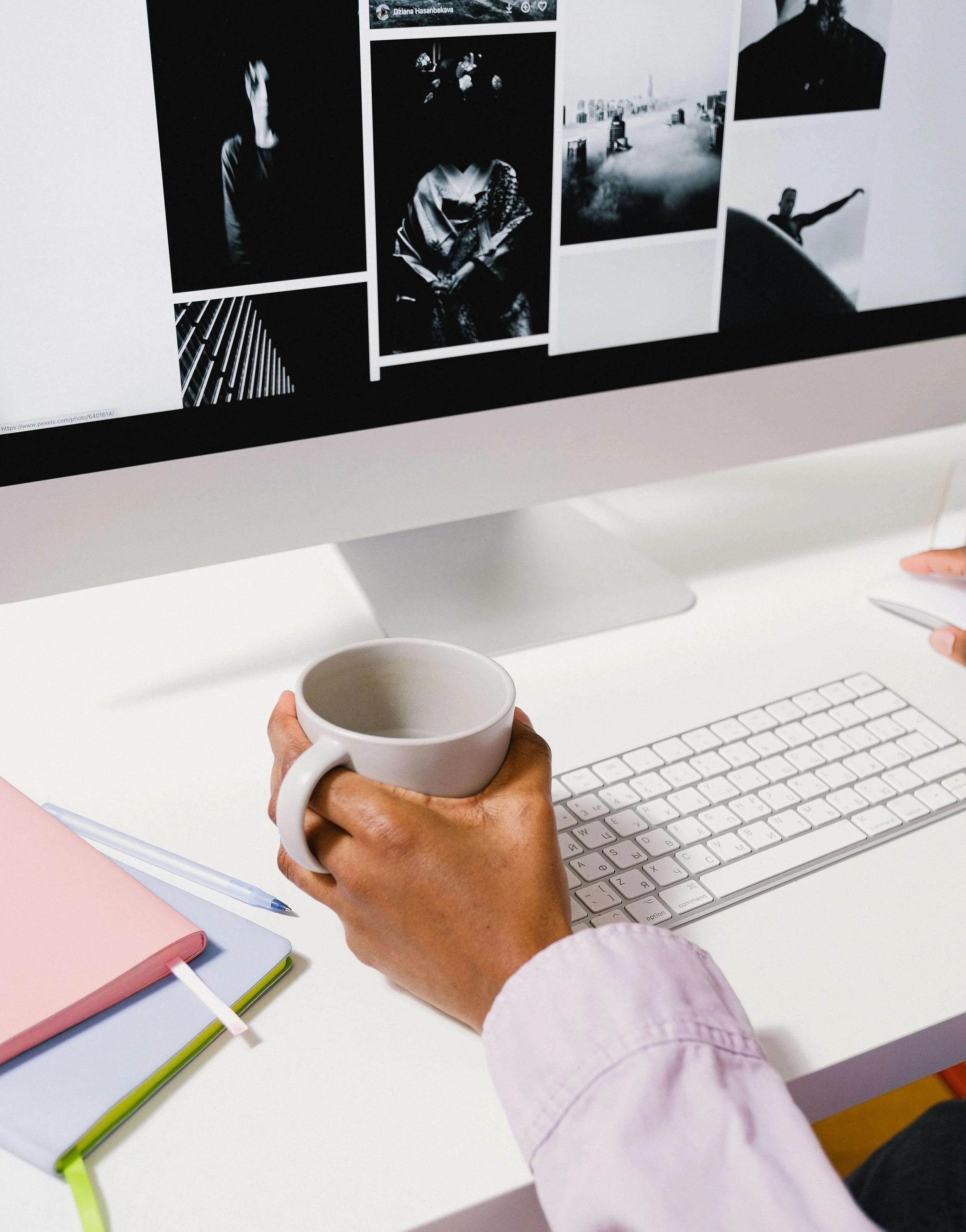 Person holding a white coffee mug and working at a white desk with a computer, notebooks, and a pen, viewing black and white photos on the computer screen.