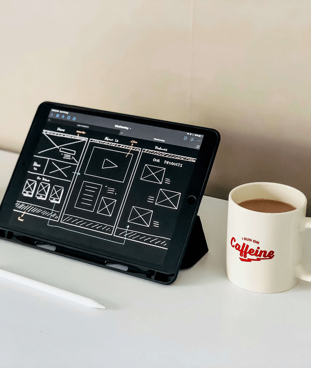 A tablet displaying a wireframe of a website, placed on a white surface next to a white mug with red text that says "I run on Caffeine," filled with a hot beverage.