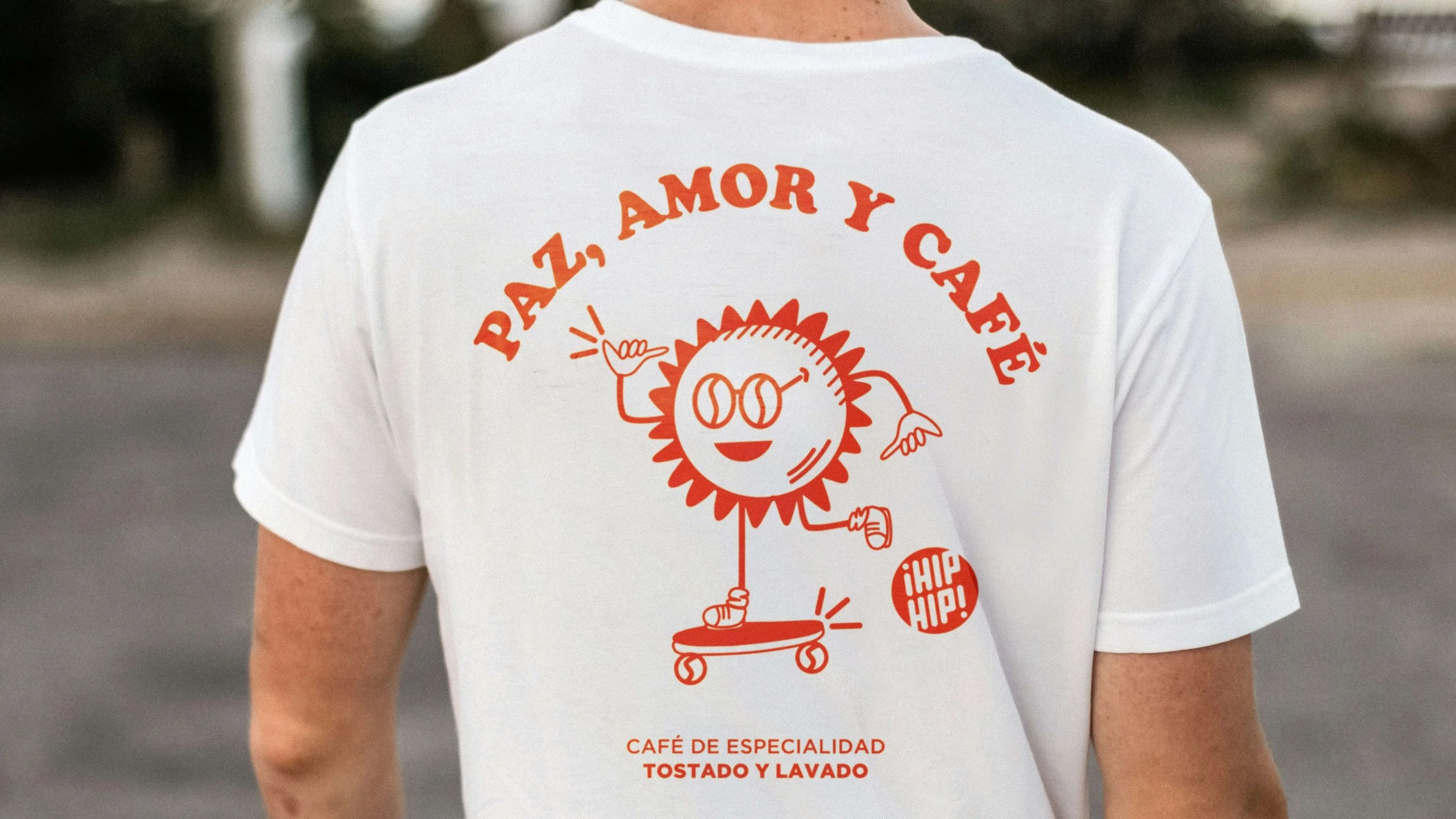 Back of a white t-shirt with orange graphic design featuring a smiling sun character with sunglasses, skateboarding, and text in Spanish that reads 'Paz, Amor y Café'. Additional text says 'Café de Especialidad Tostado y Lavado' and a small red circle with '¡Hip Hip!'.