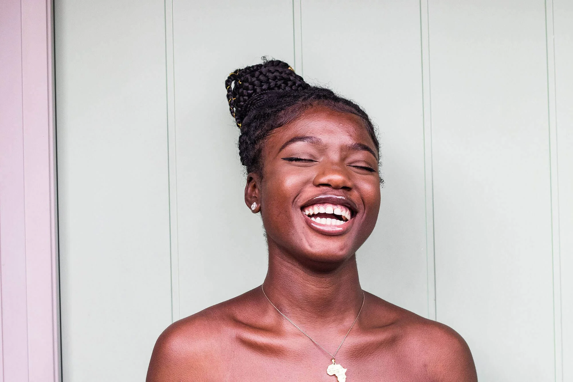 A young Black woman with braided hair, smiling with her eyes closed, wearing a necklace with the shape of Africa, standing against a pale green wall.