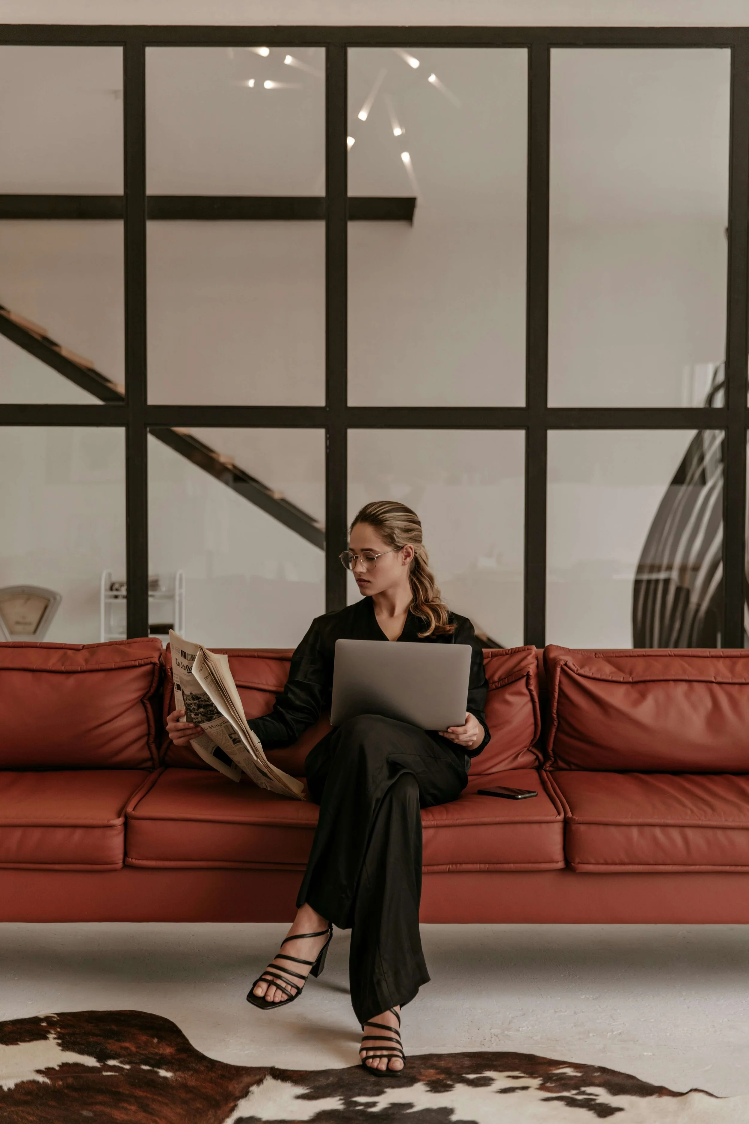 A woman dressed in black sitting on a red couch, reading a newspaper in one hand and holding a laptop on her lap, with a smartphone beside her on the cushion. She is wearing glasses and high heel sandals, with a modern interior background featuring large window panes and a staircase.