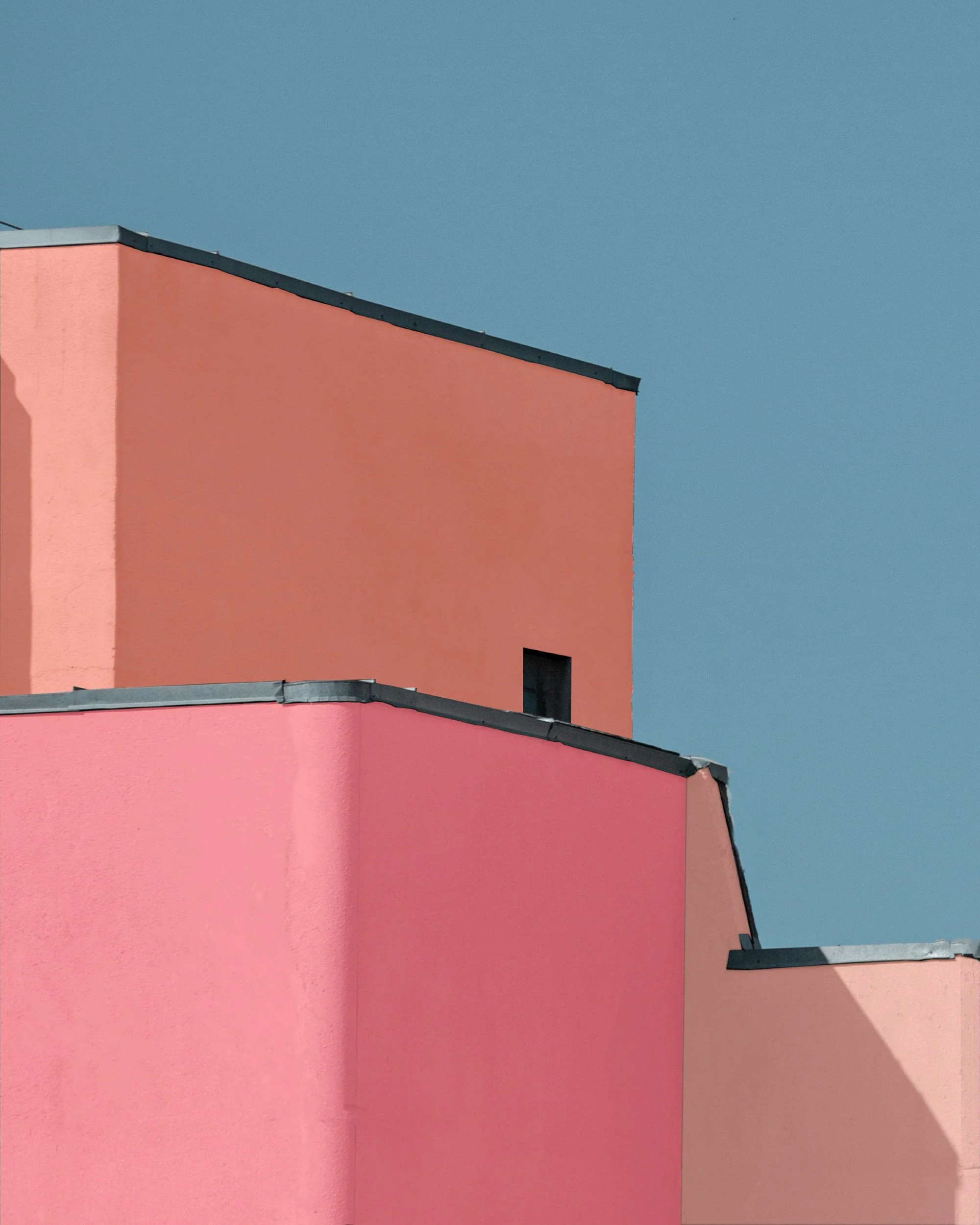 Close-up of colorful geometric building walls in pink, coral, and peach, with dark roofing edges against a blue sky.
