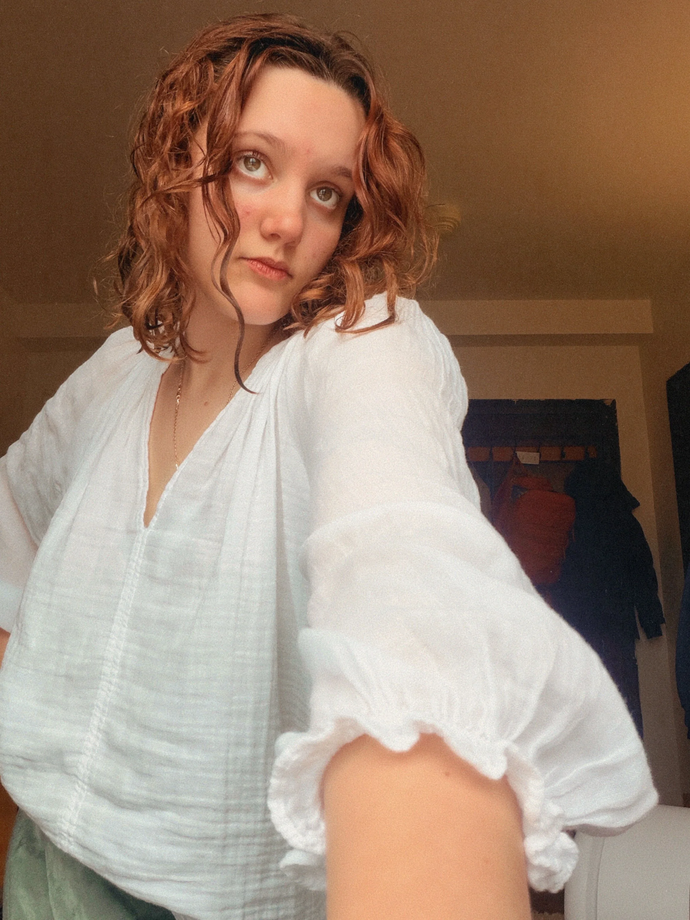 A woman with curly auburn hair, wearing a white blouse, taking a selfie in a room.