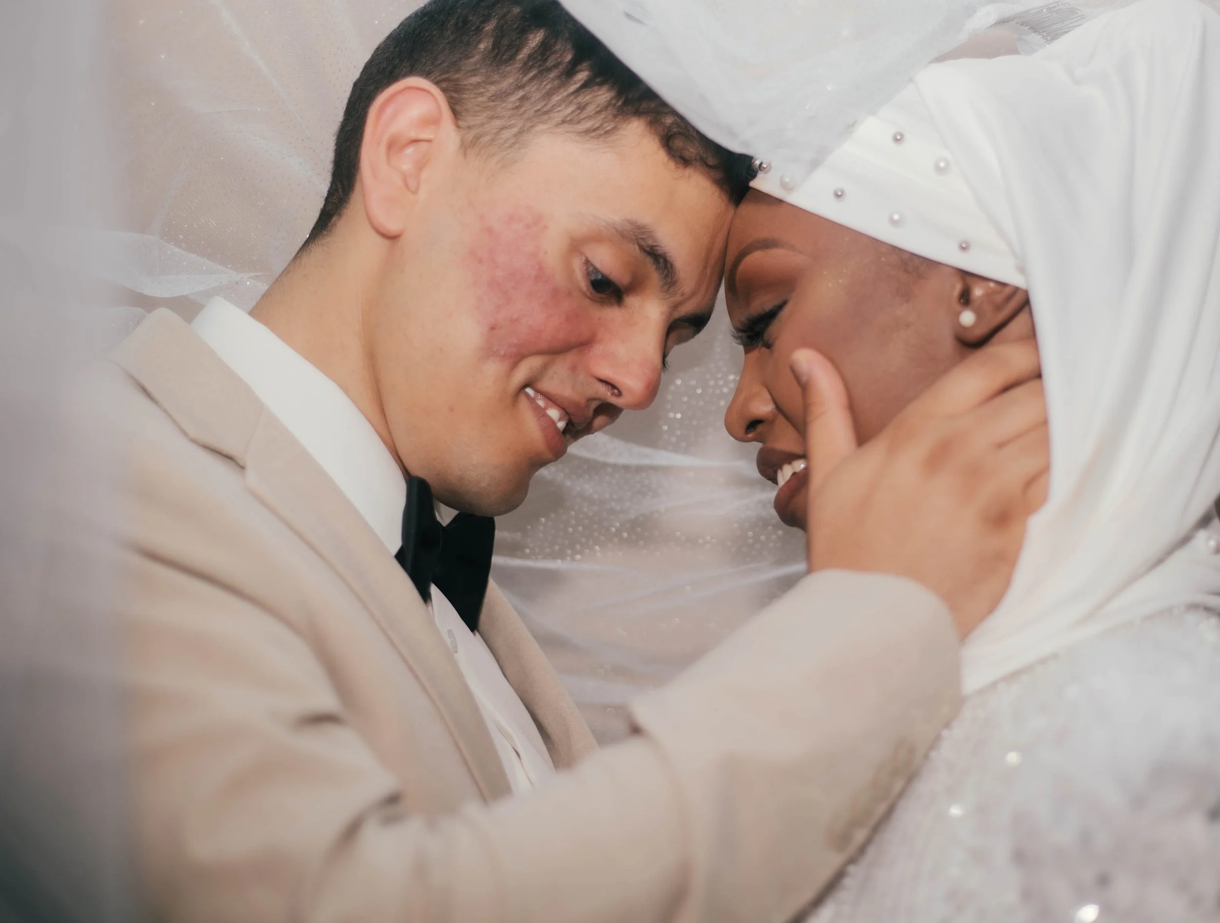 Close-up of a smiling couple, a man and a woman, with foreheads touching and eyes closed, dressed in formal attire, in an intimate moment on their wedding day.