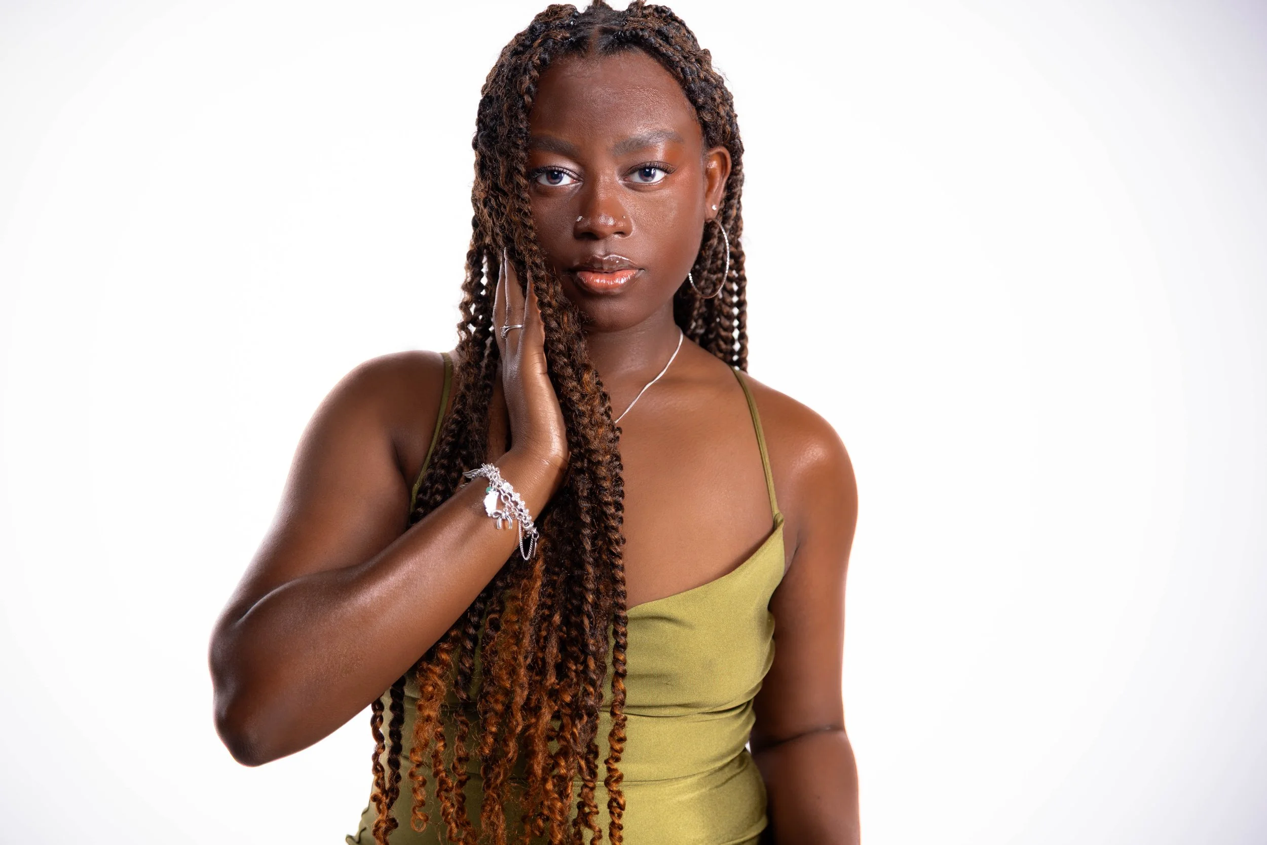 A young woman with long, twisted hair wearing a green spaghetti strap top, jewelry, and hoop earrings, touching her face with her left hand, against a plain white background.