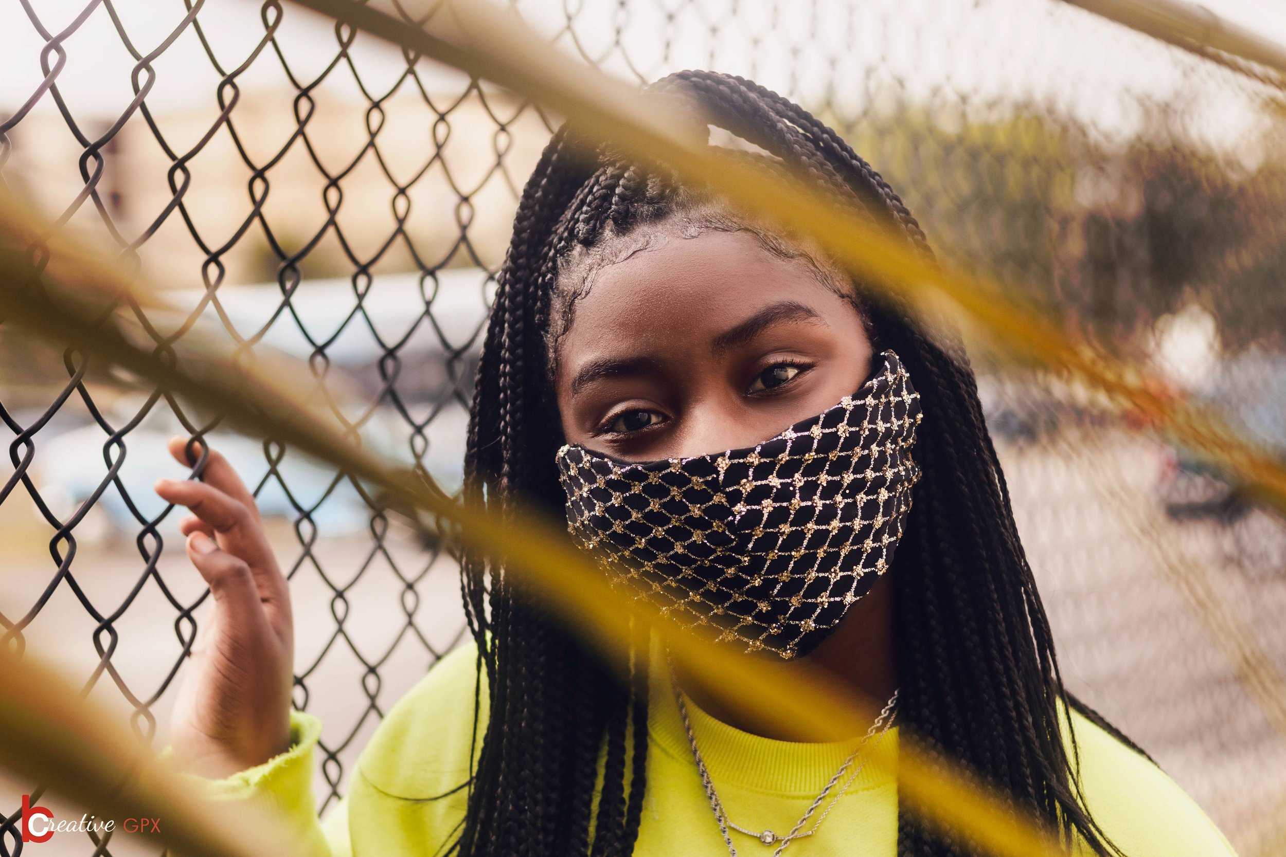 A young woman with braided hair and dark skin, wearing a patterned face mask and a yellow sweatshirt, standing behind a chain-link fence and holding onto it with her hand, outdoors.