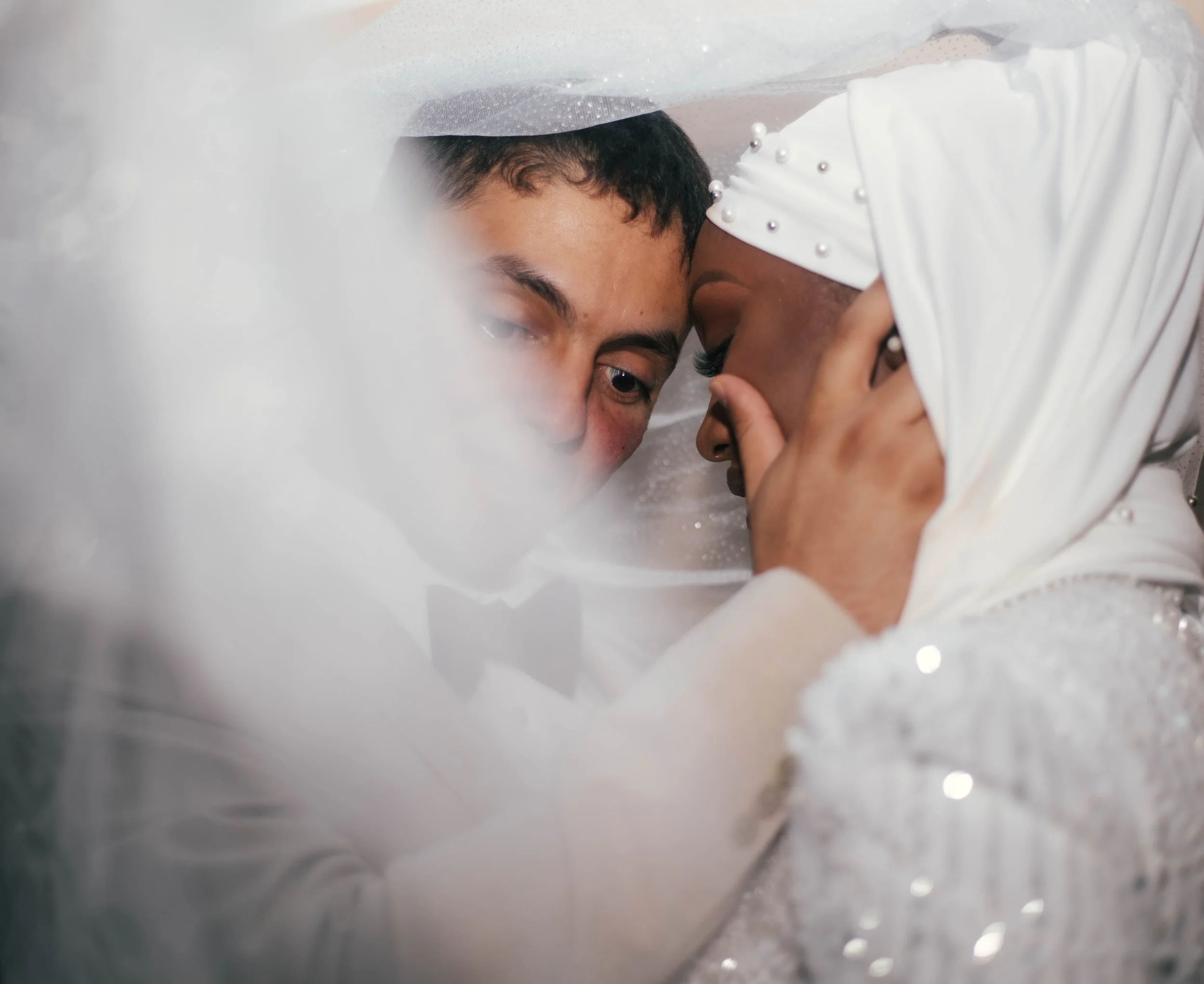 A bride and groom sharing a close, intimate moment during their wedding, with faces touching and eyes closed, surrounded by a soft, dreamy veil and sparkly details on her dress.