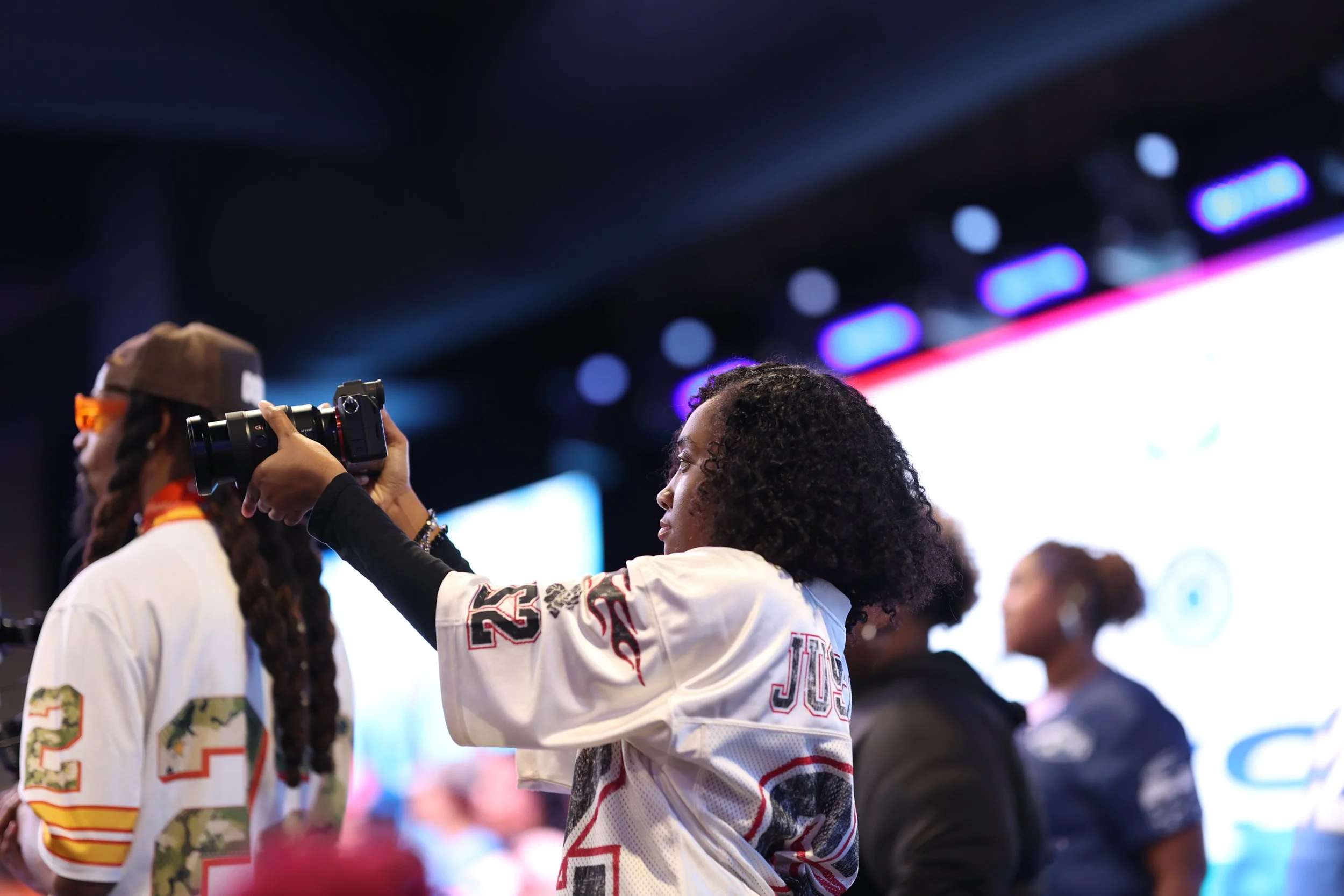 A woman taking a photo with a camera at an indoor event with multiple people in the background.