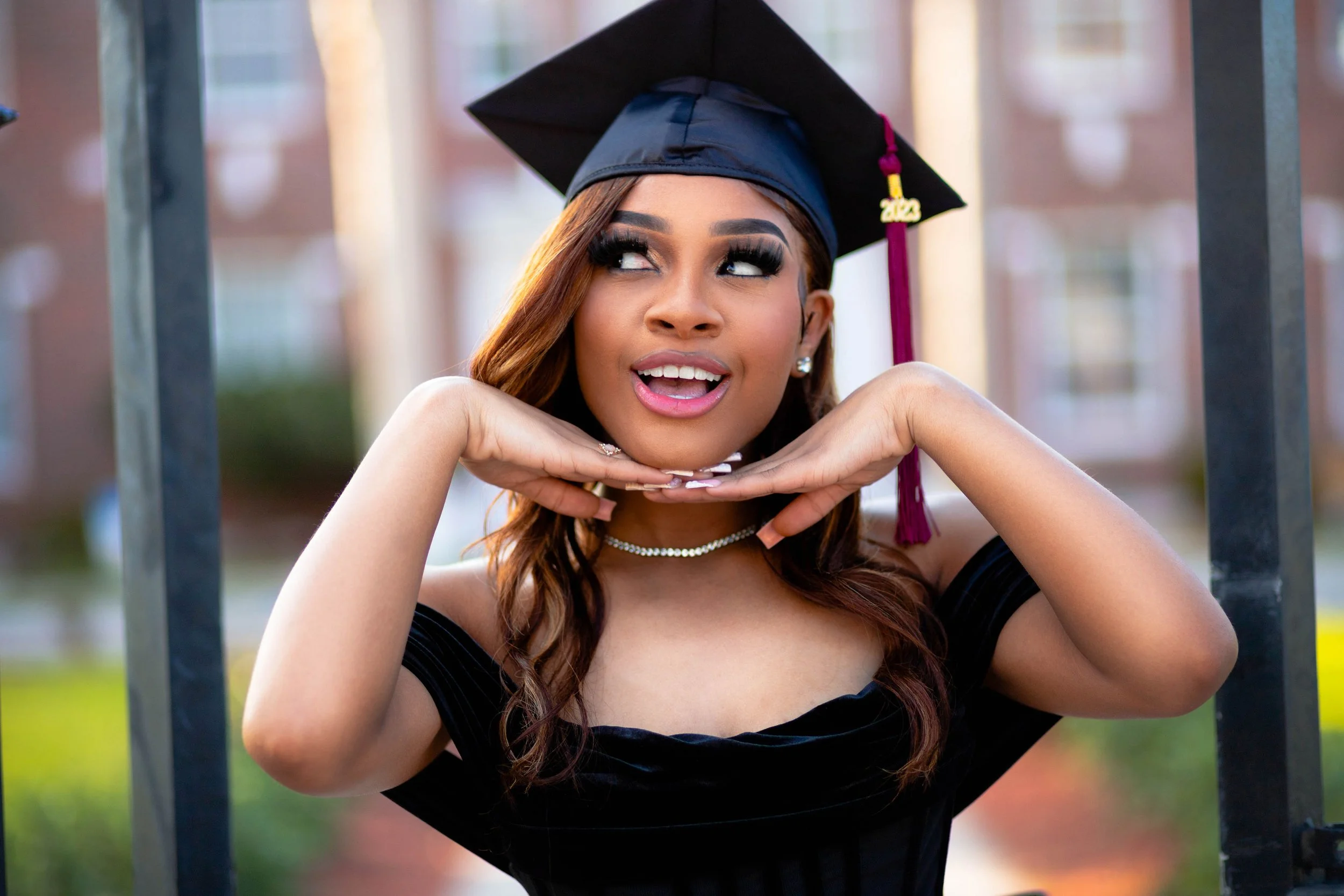Young woman in a graduation cap and gown posing outdoors, resting her chin on her hands, smiling and looking to the side.
