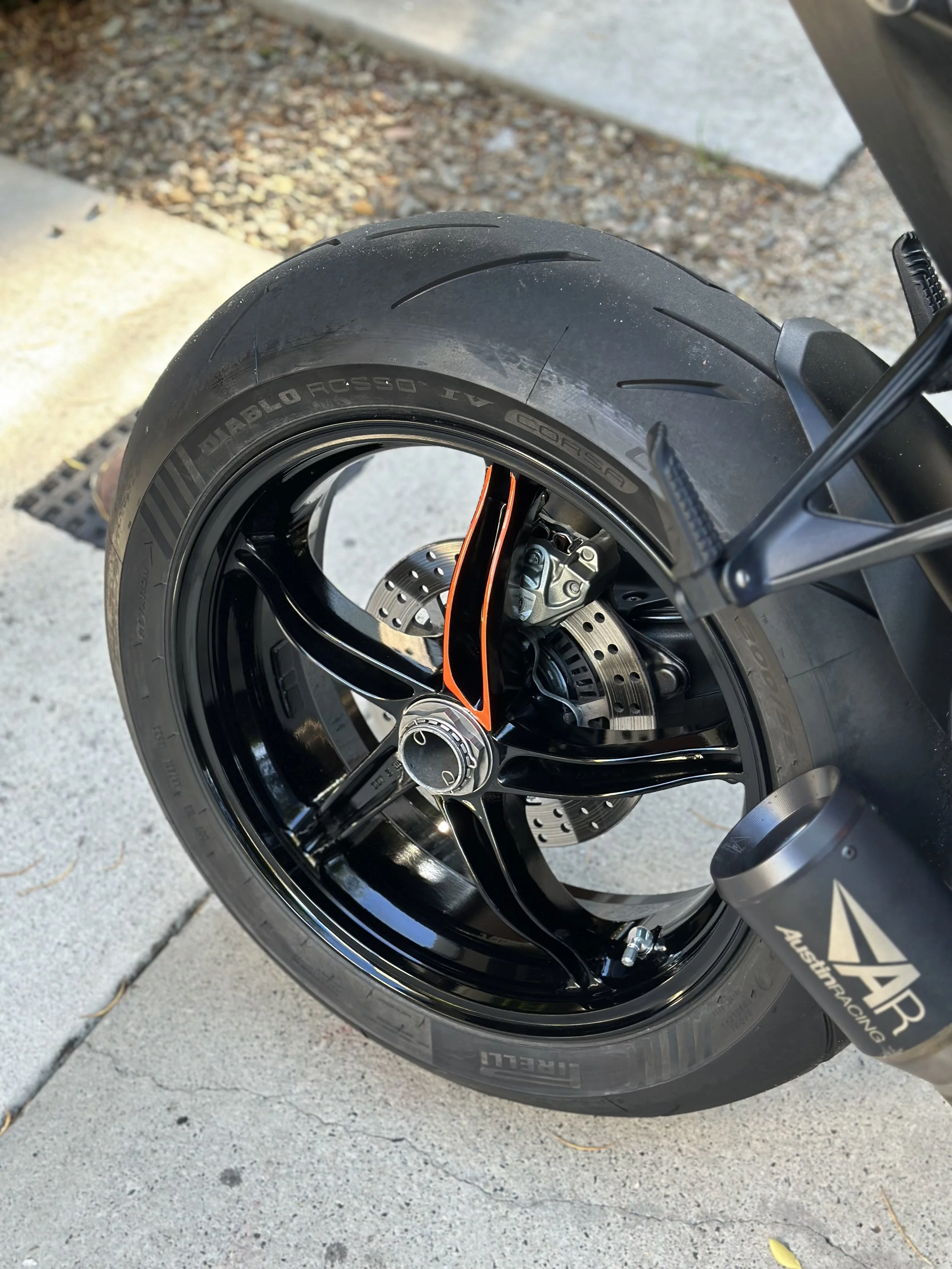 Close-up of a motorcycle rear wheel, tire, black wheel rim, disc brake, and exhaust pipe with 'Austrian Racing' branding.