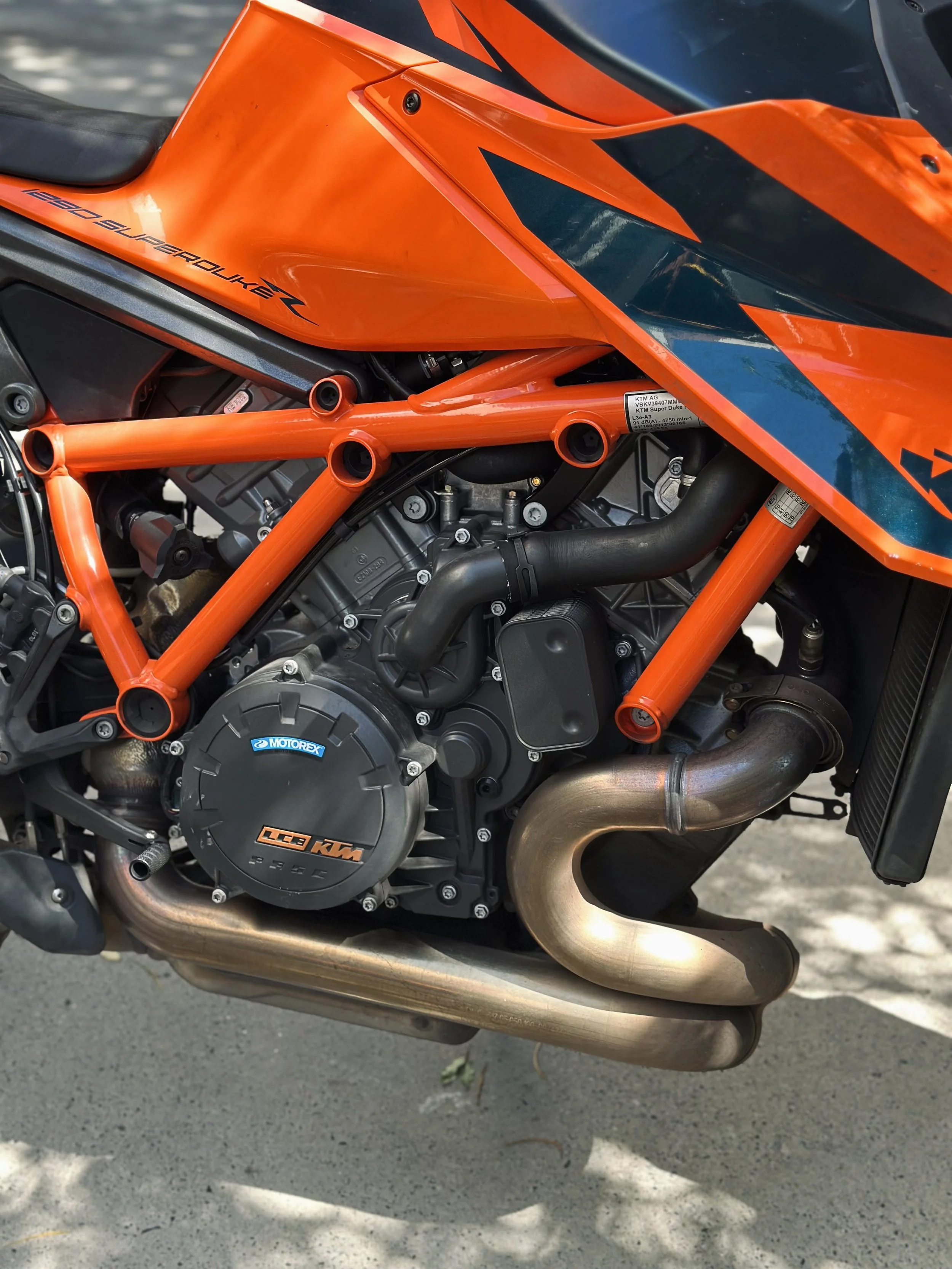 Close-up of a KTM motorcycle engine with orange frame and black and orange bodywork. The engine has a black cover with KTM branding, and the exhaust pipe is visible, made of metal.