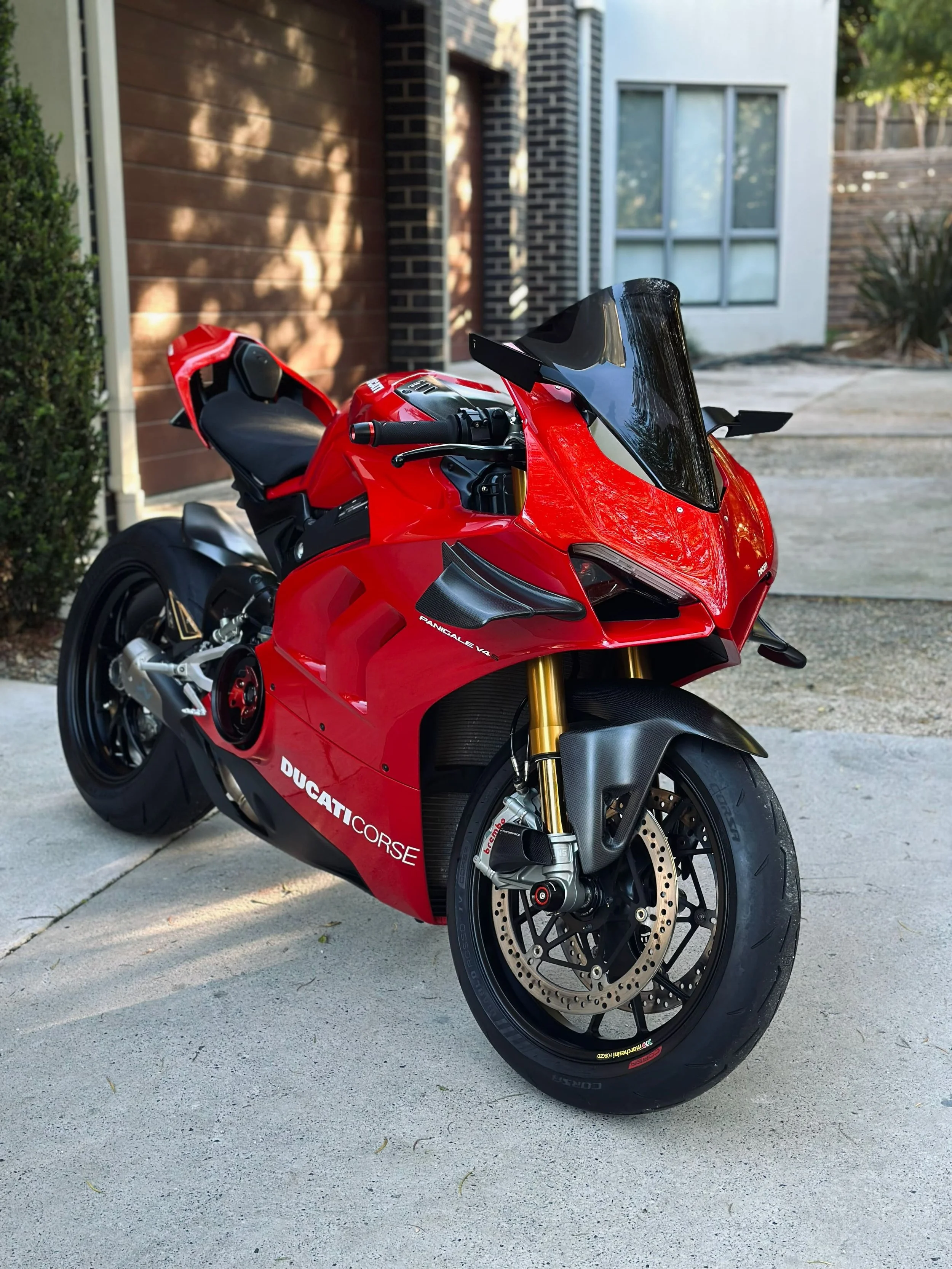 A red Ducati Corse Panigale V4 motorcycle parked outdoors on a concrete driveway, with a residential background including a brown brick wall, a wooden fence, and a house with large windows.