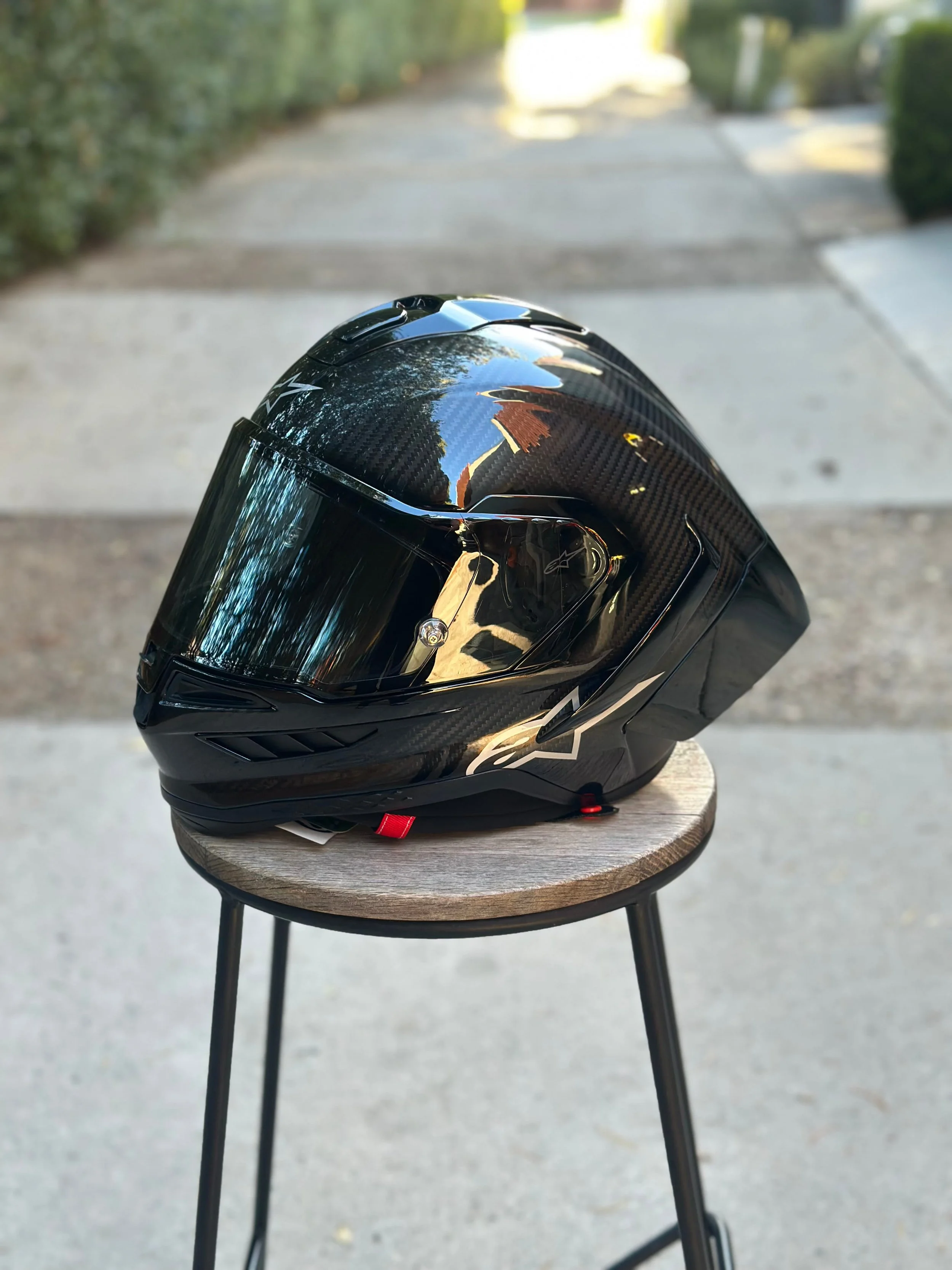 Black motorcycle helmet with a dark visor, placed on a small round wooden stool outside on a concrete surface, with trees and a sidewalk in the background.
