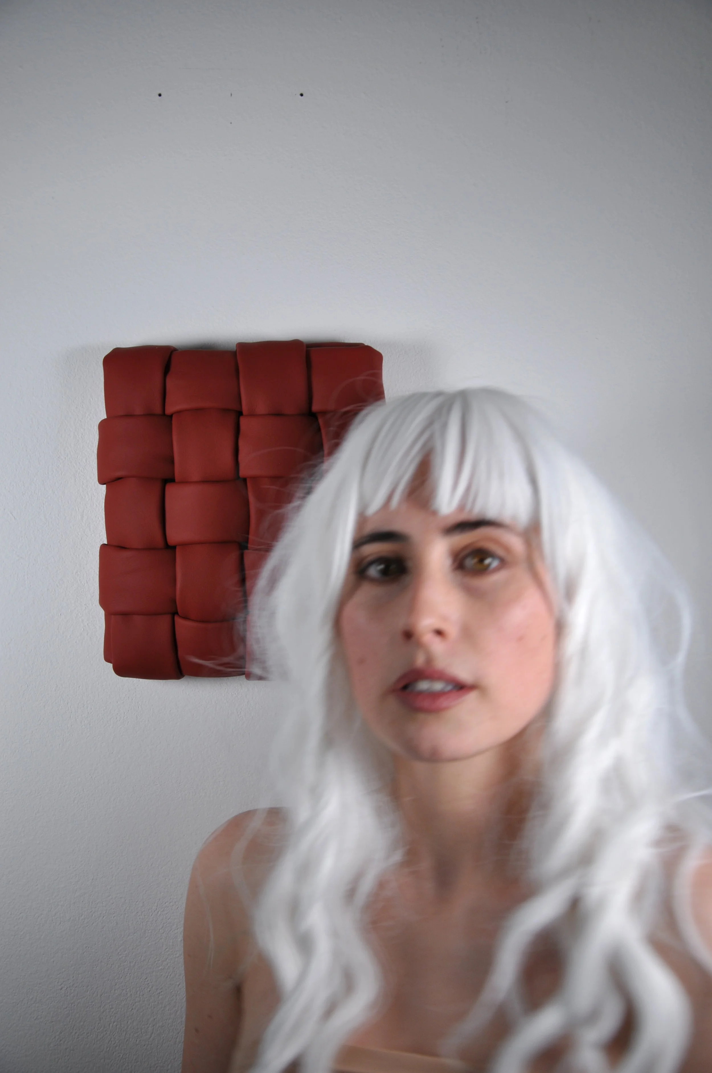 Close-up of a woman with white hair, standing against a plain wall with a dark red art work. The photo captures her face and upper shoulders. The textile artwork is made out of padded leather woven into a fine grid weave.