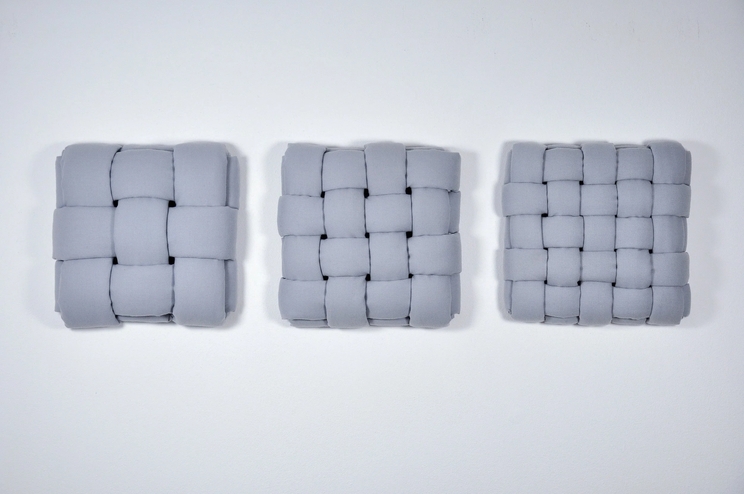 Three square, light gray woven textile artworks mounted horizontally in a row against a plain white wall. From left to right each piece features a progressively finer grid weave, constructed using thick, padded fabric tubes woven in 3, 4 & 5 strands.