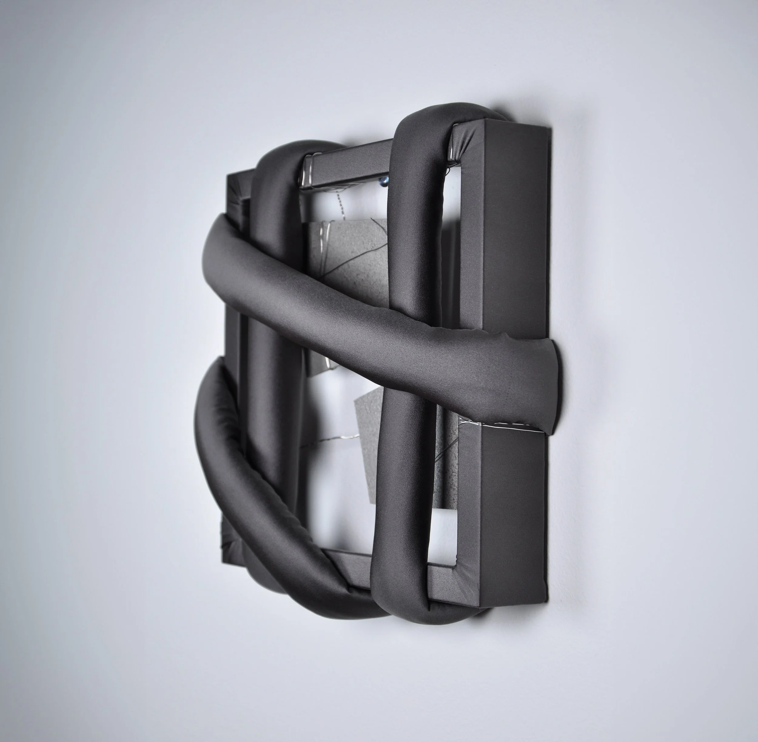 Abstract textile art piece featuring thick, dark grey padded fabric tubes loosely woven around a matching fabric-wrapped square frame, revealing grey stone-like plates bound with silver wire in the center.
