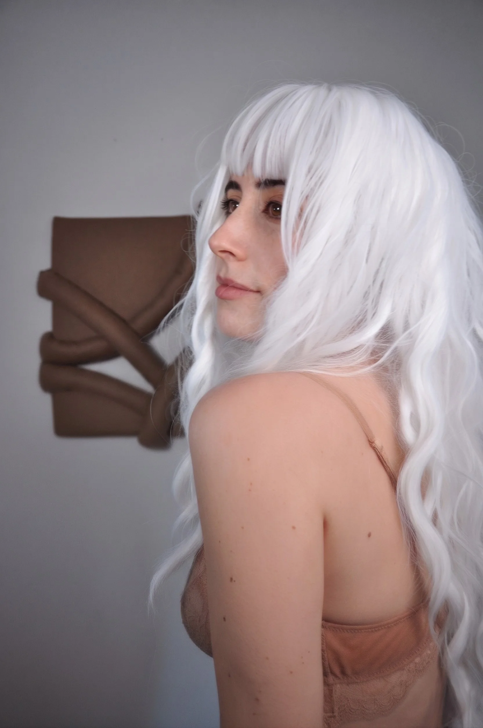 Profile of a woman with long white hair, wearing a nude colored top. In the background you see a textile artwork against a white wall. The fabric art is made out of a wool fabric, in a camel tone and plays with padding and negative space.