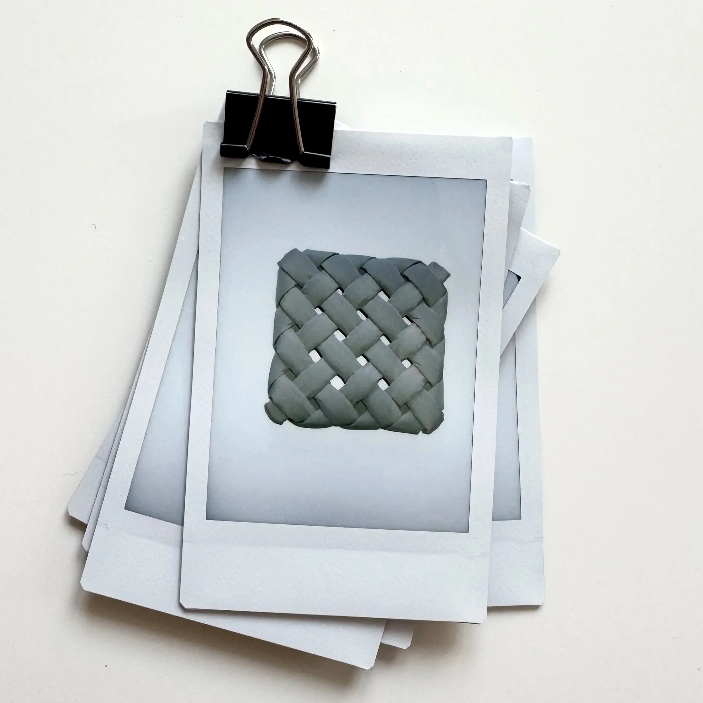 A binder clip attaching a stack of polaroid photos. The photo on top shows a sand colored woven textile art piece against a plain white wall.  It is a very modern picture of an analoge archive of textile art works.