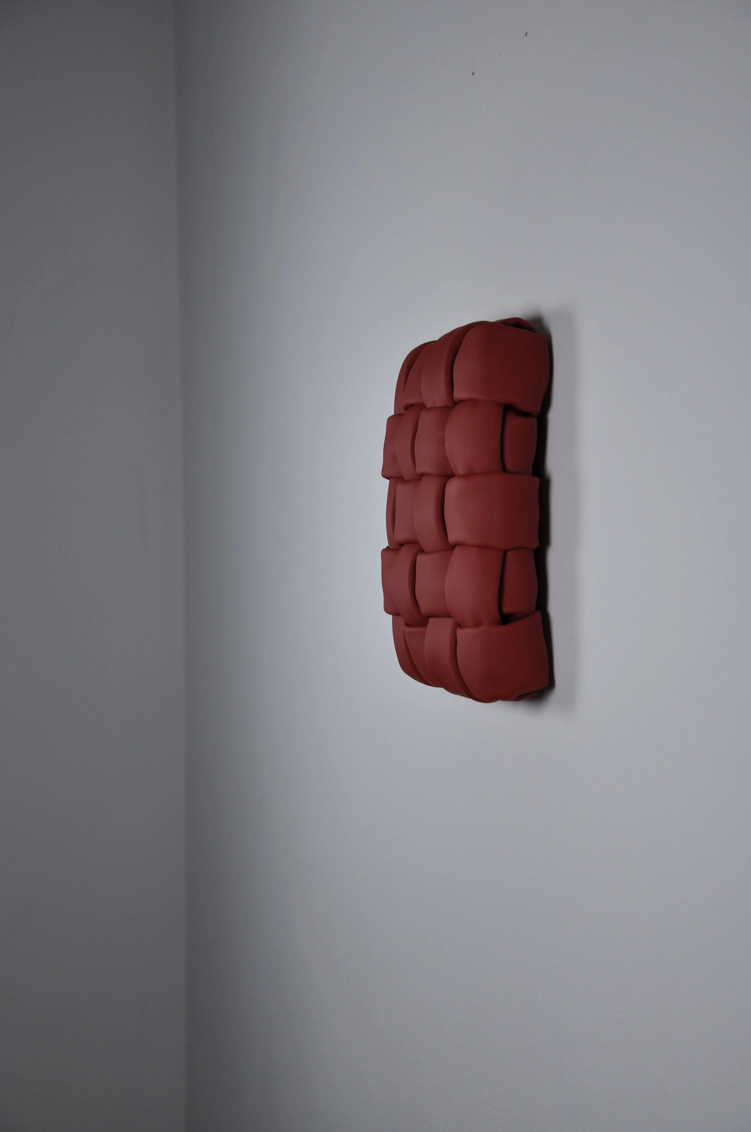 A red fabric art piece attached to a white wall. The art work is made of dark red leather. It is padded and woven into a composition of 4 vertical and 5 horizontal strings.