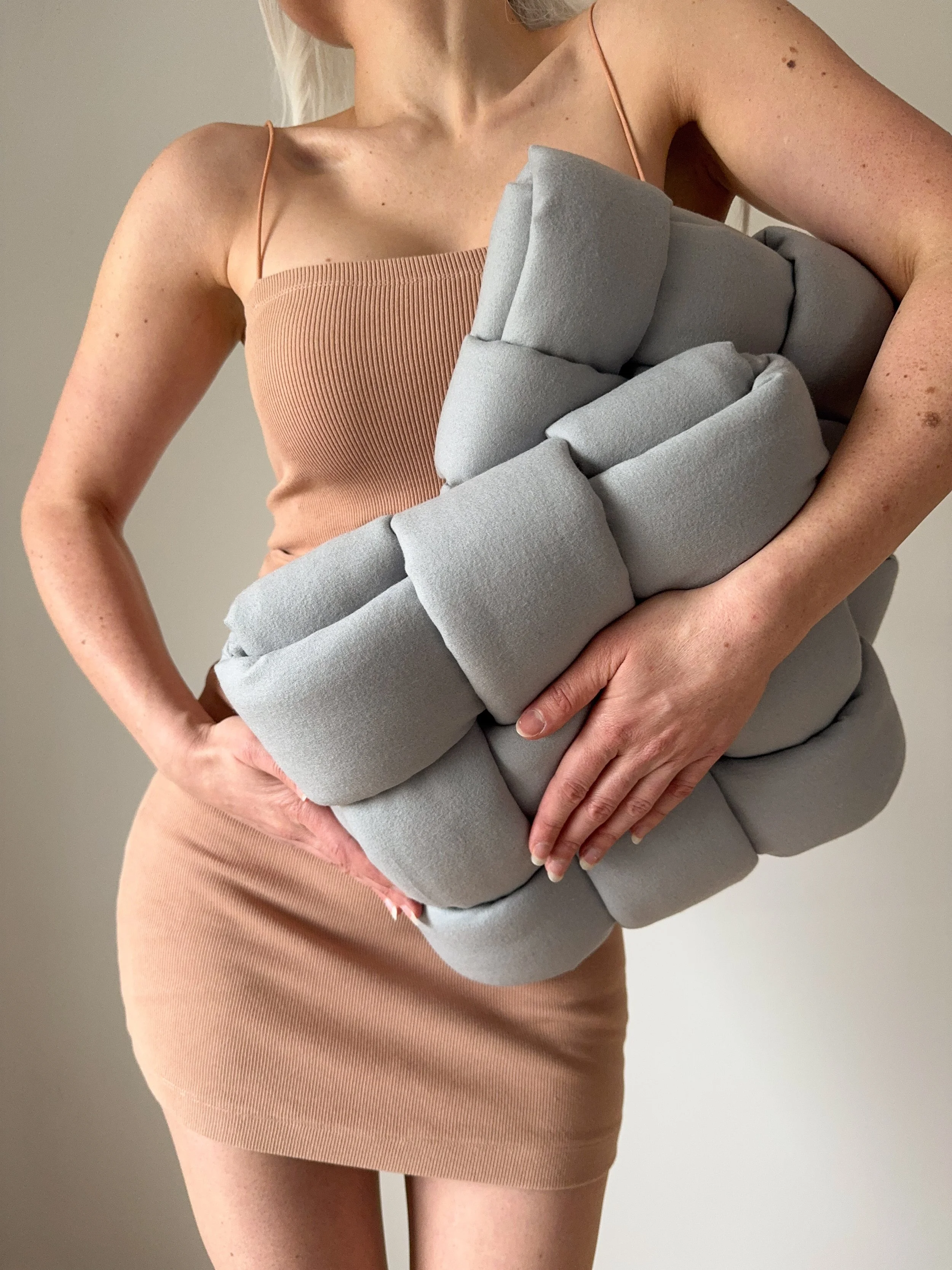 A woman in a nude dress, with white hair holding a gray padded textile art work with her hands. The art piece is made out of padded, soft wool tubes that are woven into a clean pattern. It is a three-dimensional textile art piece.