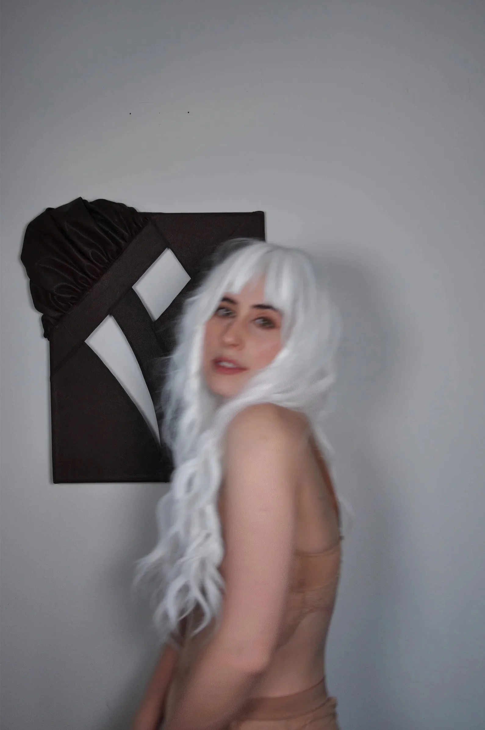 A woman with long white hair and light skin, wearing nude colored lingerie, stands in front of a white wall with a dark brown textile art piece. It is made out of leather and plays with ruffles, weaving and the use of negative space.