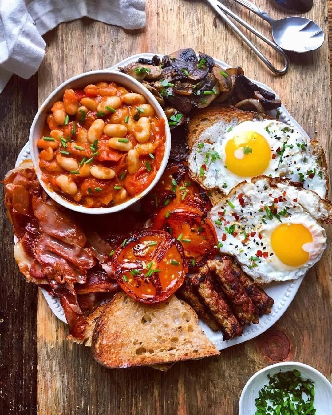 Traditional breakfast plate with baked eggs, bacon, grilled tomato, sautéed mushrooms, baked beans, toasted bread, and coffee.