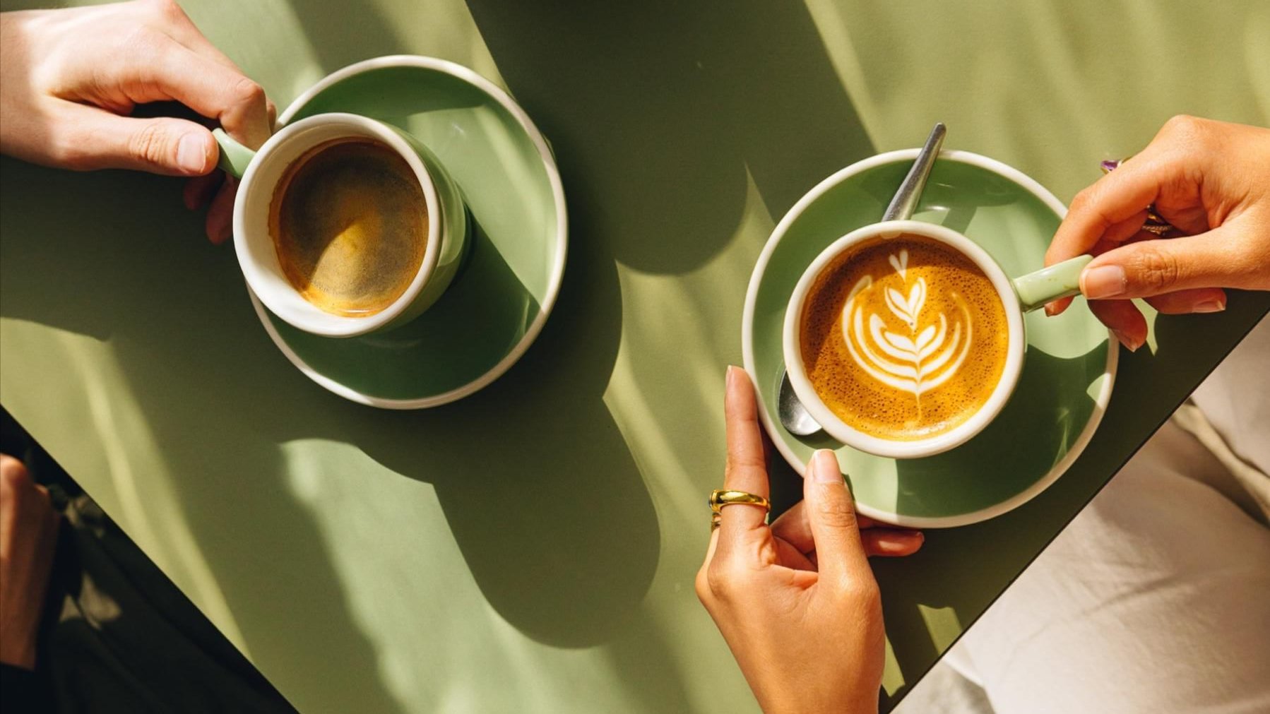 Two cups of coffee on green saucers with spoons, one with latte art and the other plain, on a green table held by two hands.