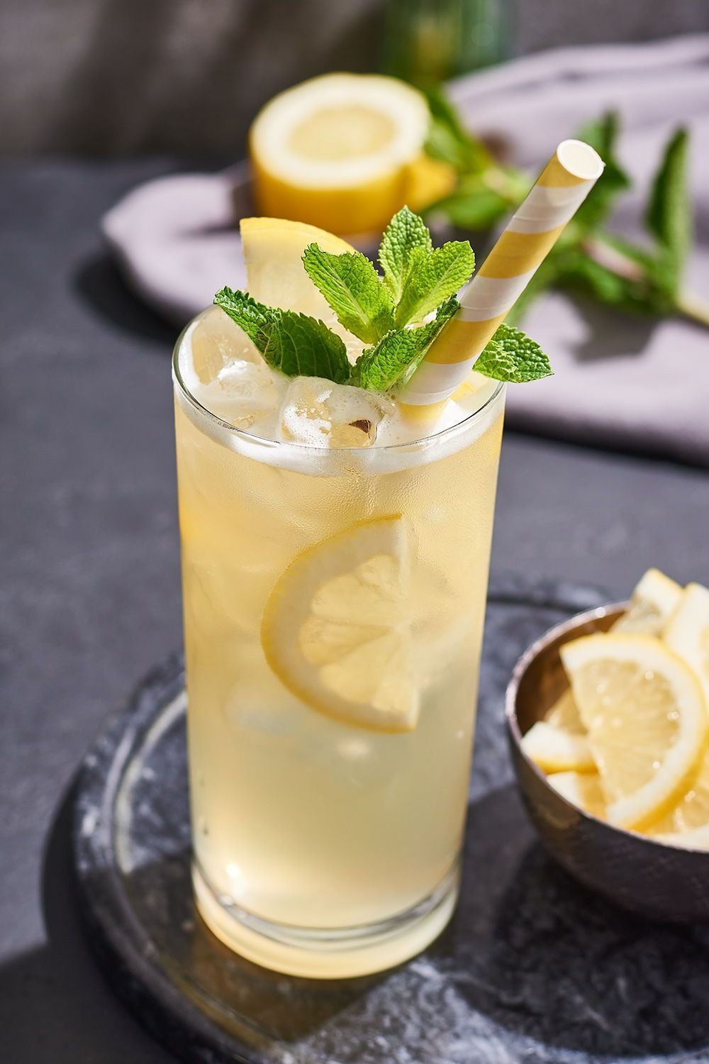 A tall glass of lemonade garnished with fresh mint leaves, lemon slices, and a striped paper straw, on a dark surface with lemon slices and a bowl of lemon wedges nearby.