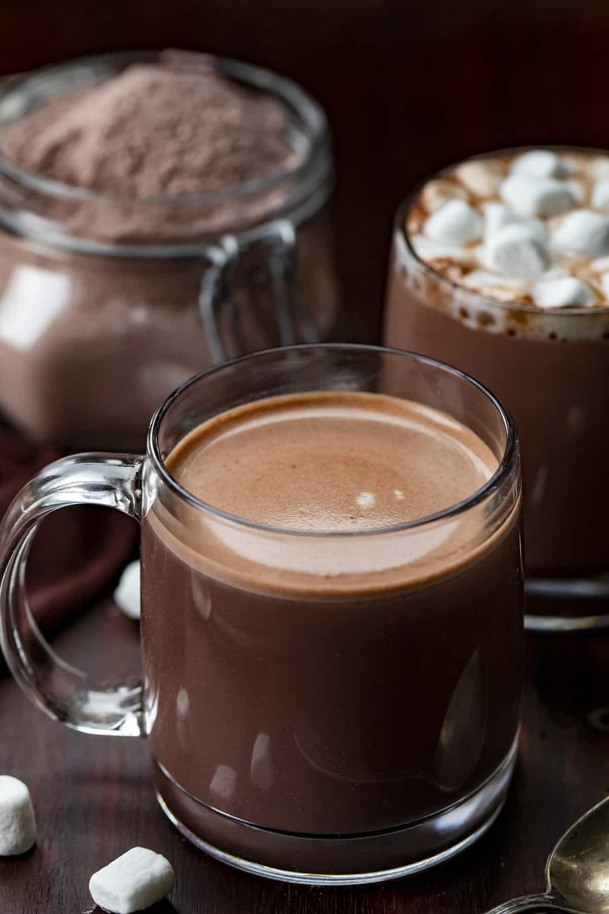 Two clear glass mugs filled with hot chocolate topped with marshmallows, placed on a wooden surface. In the background, there is a jar of cocoa powder and additional marshmallows.