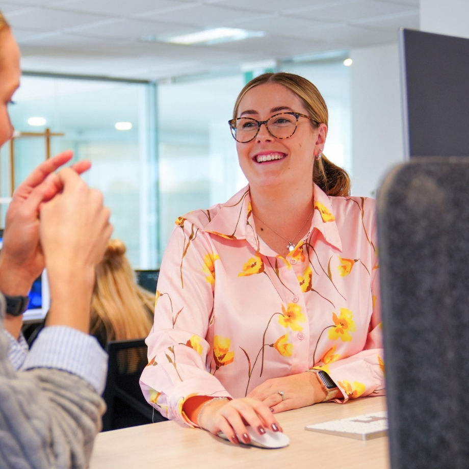 A woman wearing glasses and a pink floral blouse smiling and talking with a man in an office setting.