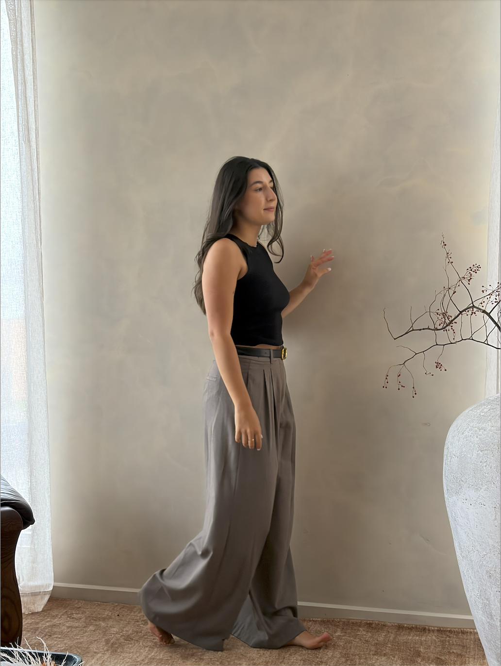 A woman with long dark hair standing barefoot in a living room, wearing a black sleeveless top and high-waisted gray wide-leg pants, touching a beige wall.