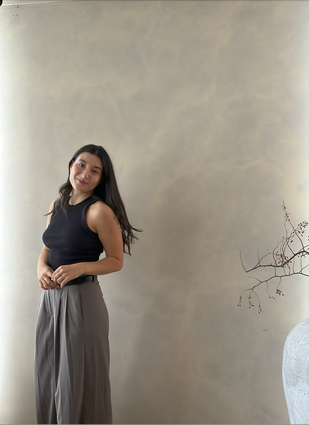 A woman with long dark hair wearing a black sleeveless top and gray wide-leg trousers, standing indoors near a beige wall and a decorative branch in a vase.